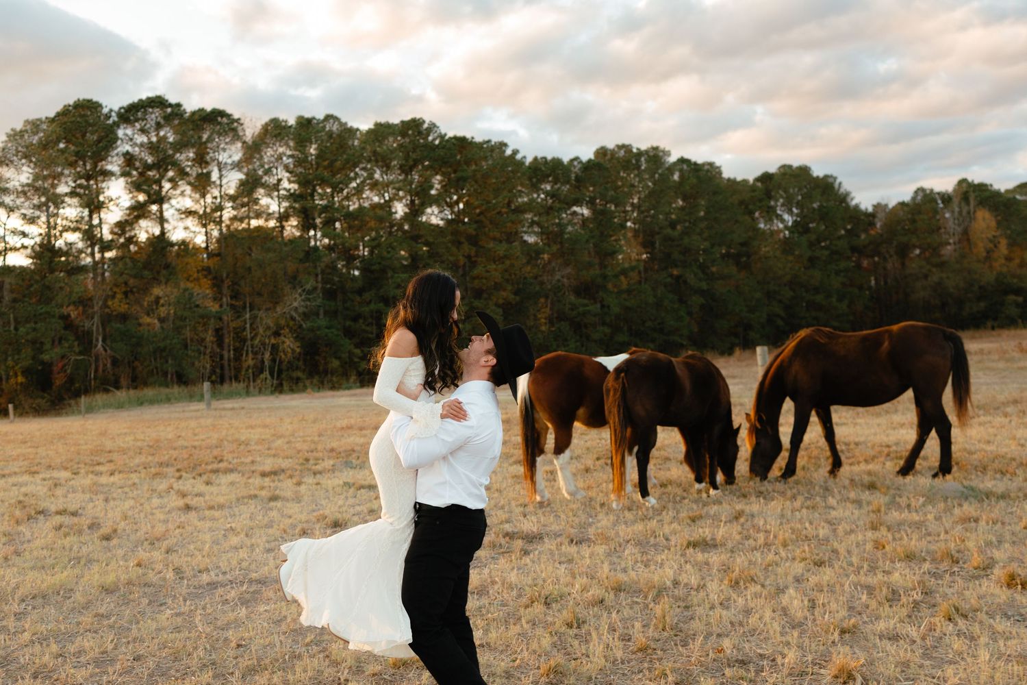 Groom twirling bride in white wedding dress in a field with three horses grazing at sunset.
