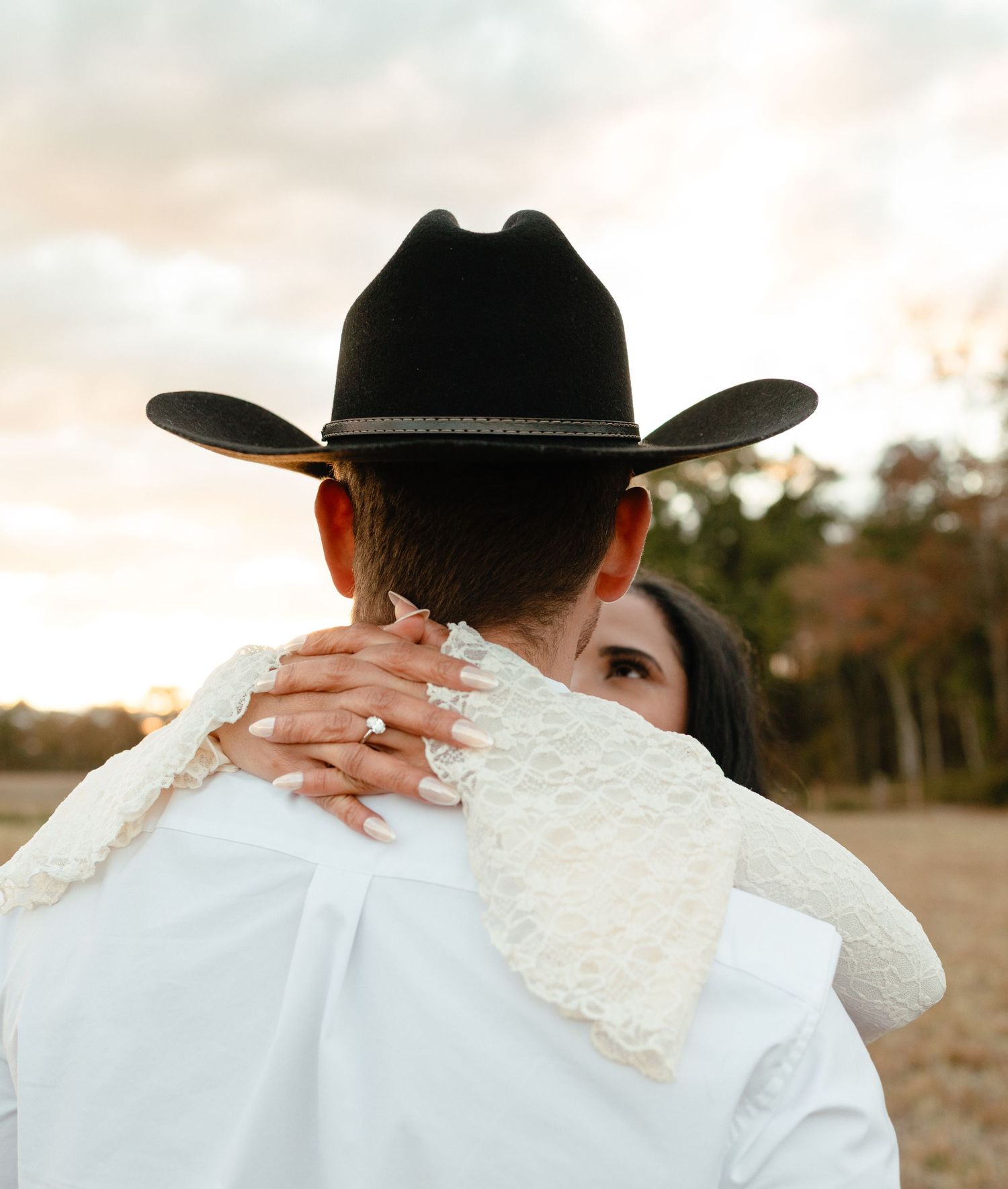 Close-up of bride embracing groom from behind, showcasing her diamond ring against his white shirt and black cowboy hat.