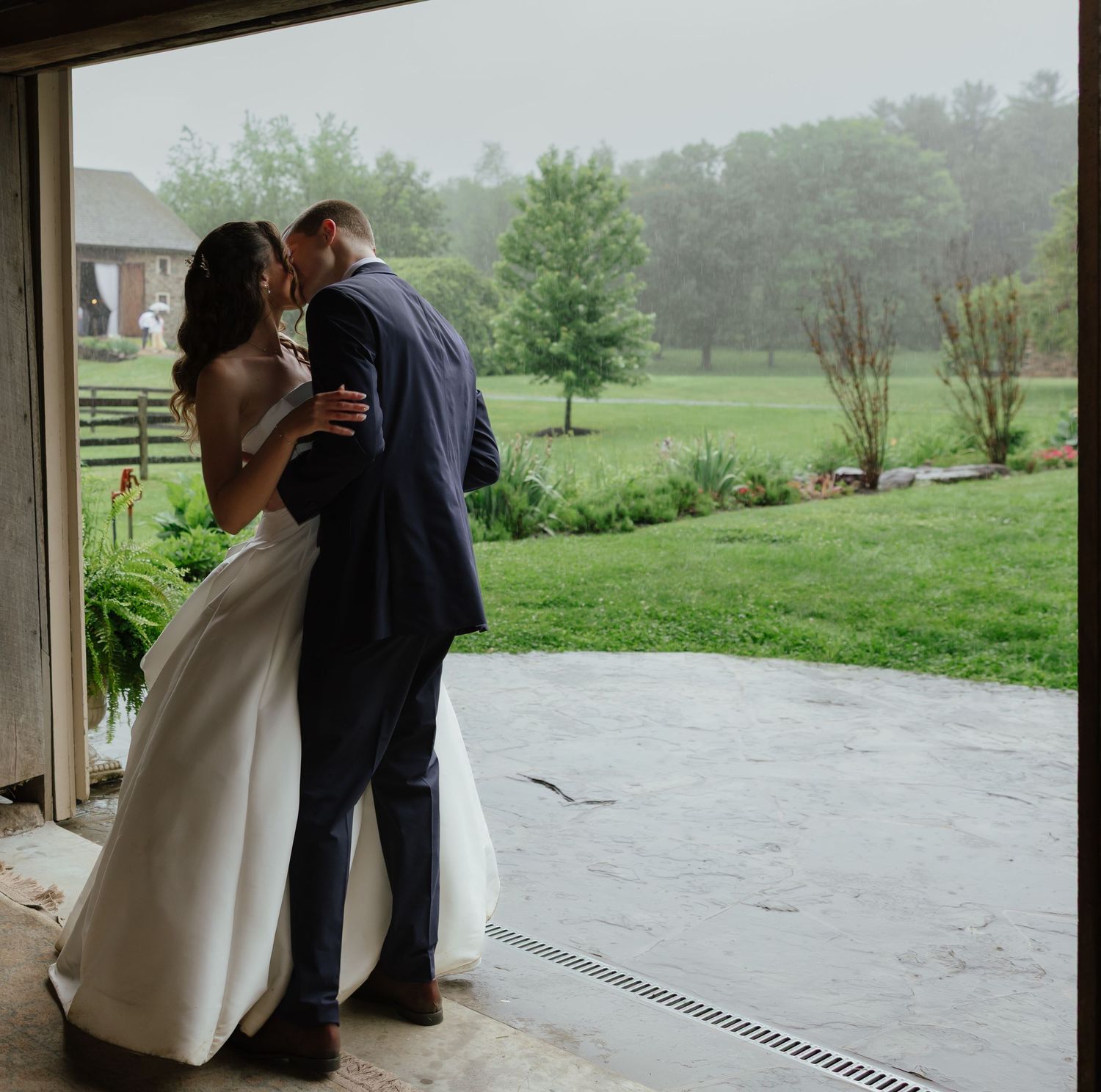 Bride and groom share a kiss in barn doorway overlooking lush green farm landscape in the rain.