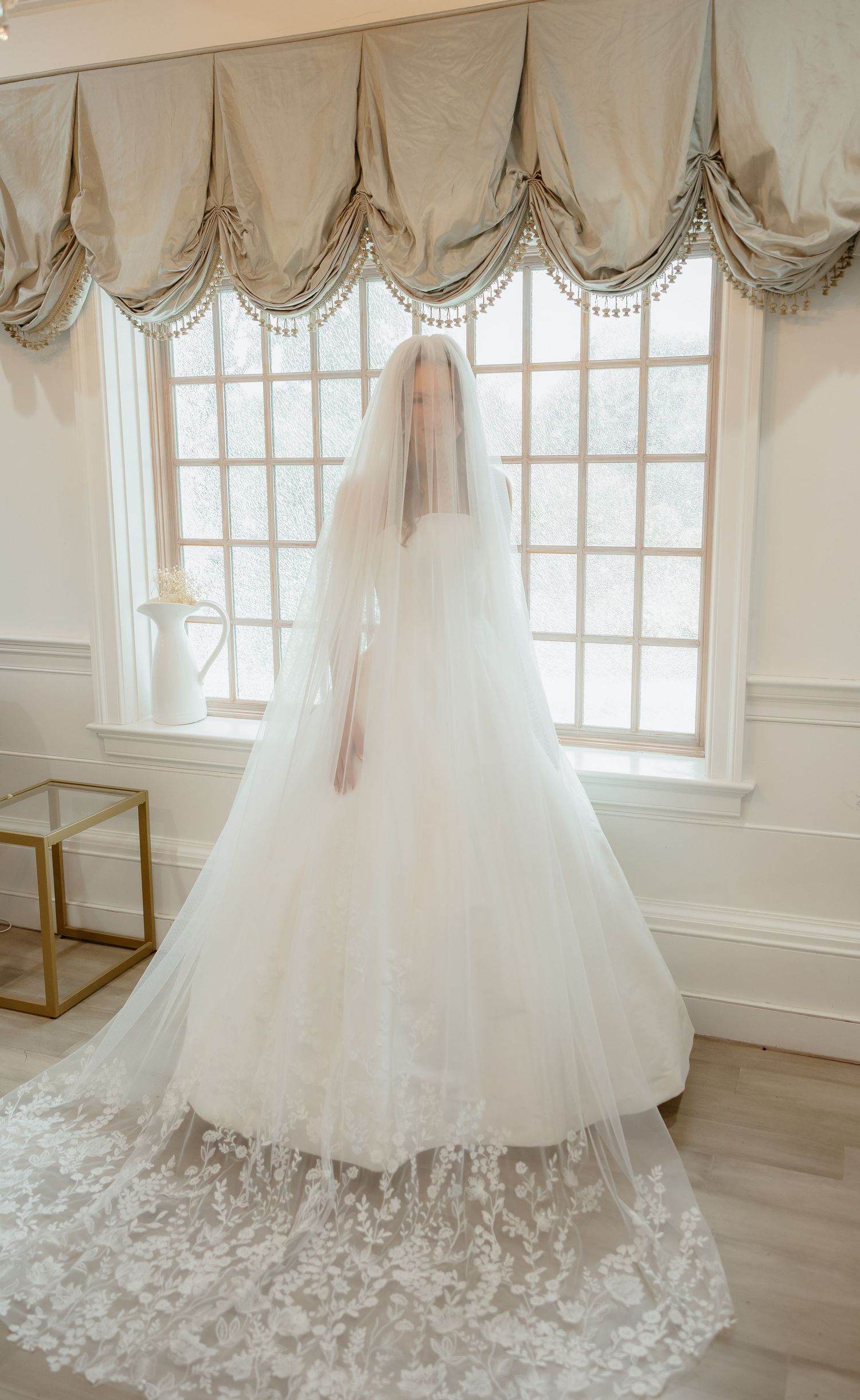Bride in a lace ball gown and cathedral veil standing before elegant draped windows.