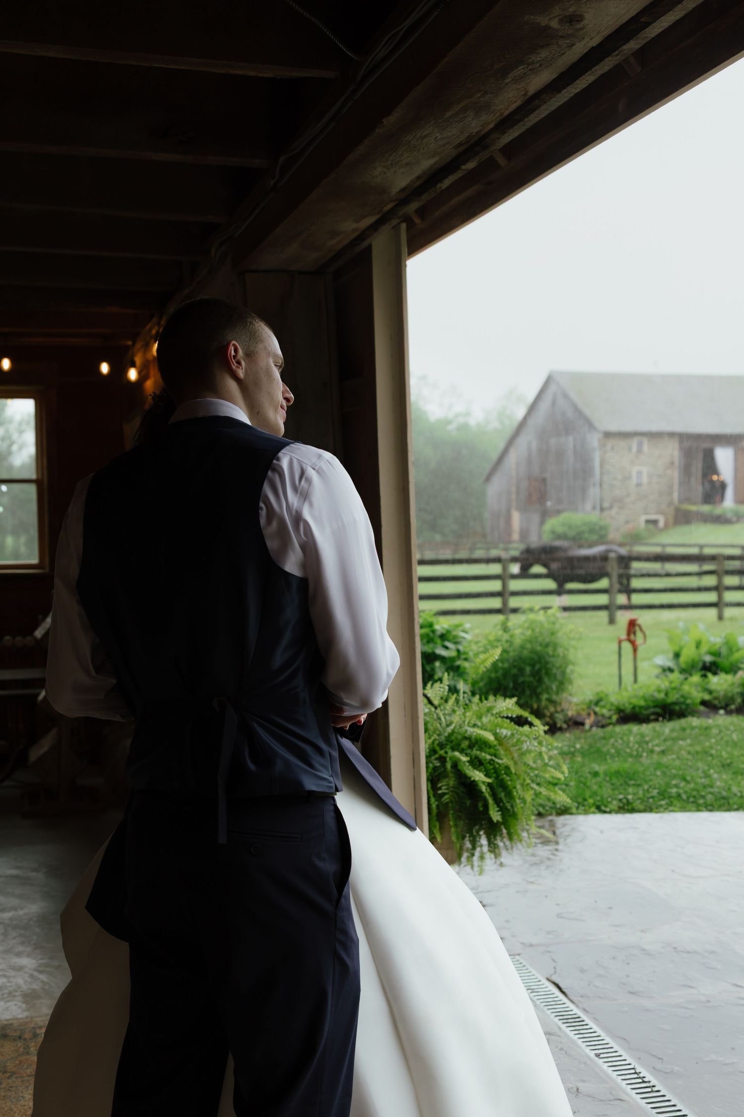 Groom in navy vest gazes out open barn door at rainy farm landscape with rustic outbuildings.