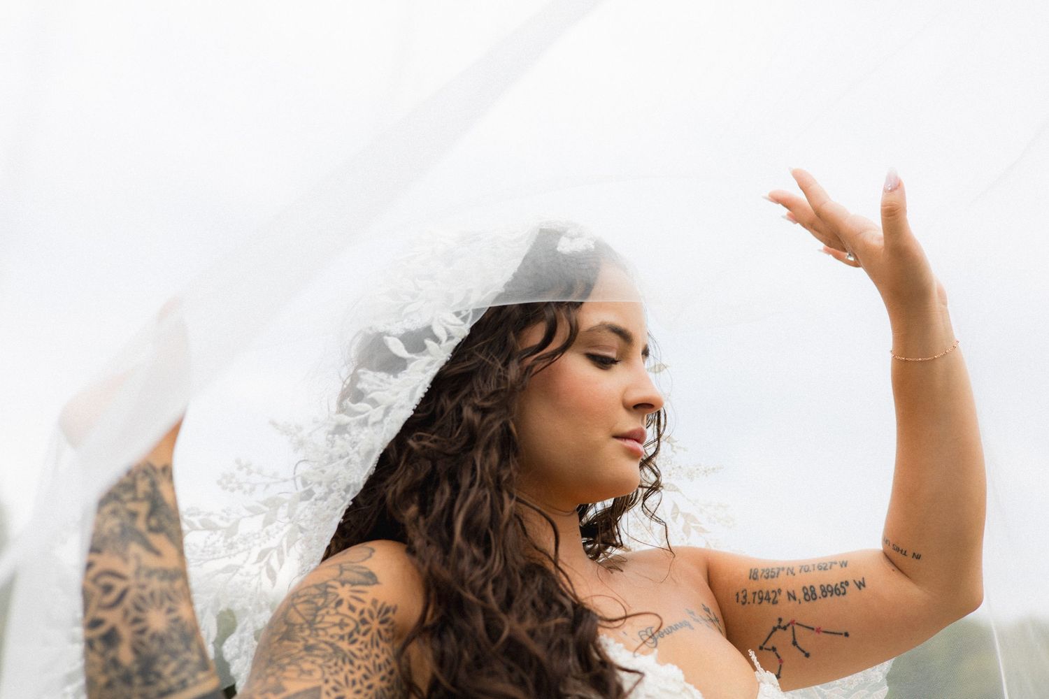 Tattooed bride in white wedding dress lifts lace veil with eyes closed in dreamy bridal portrait.
