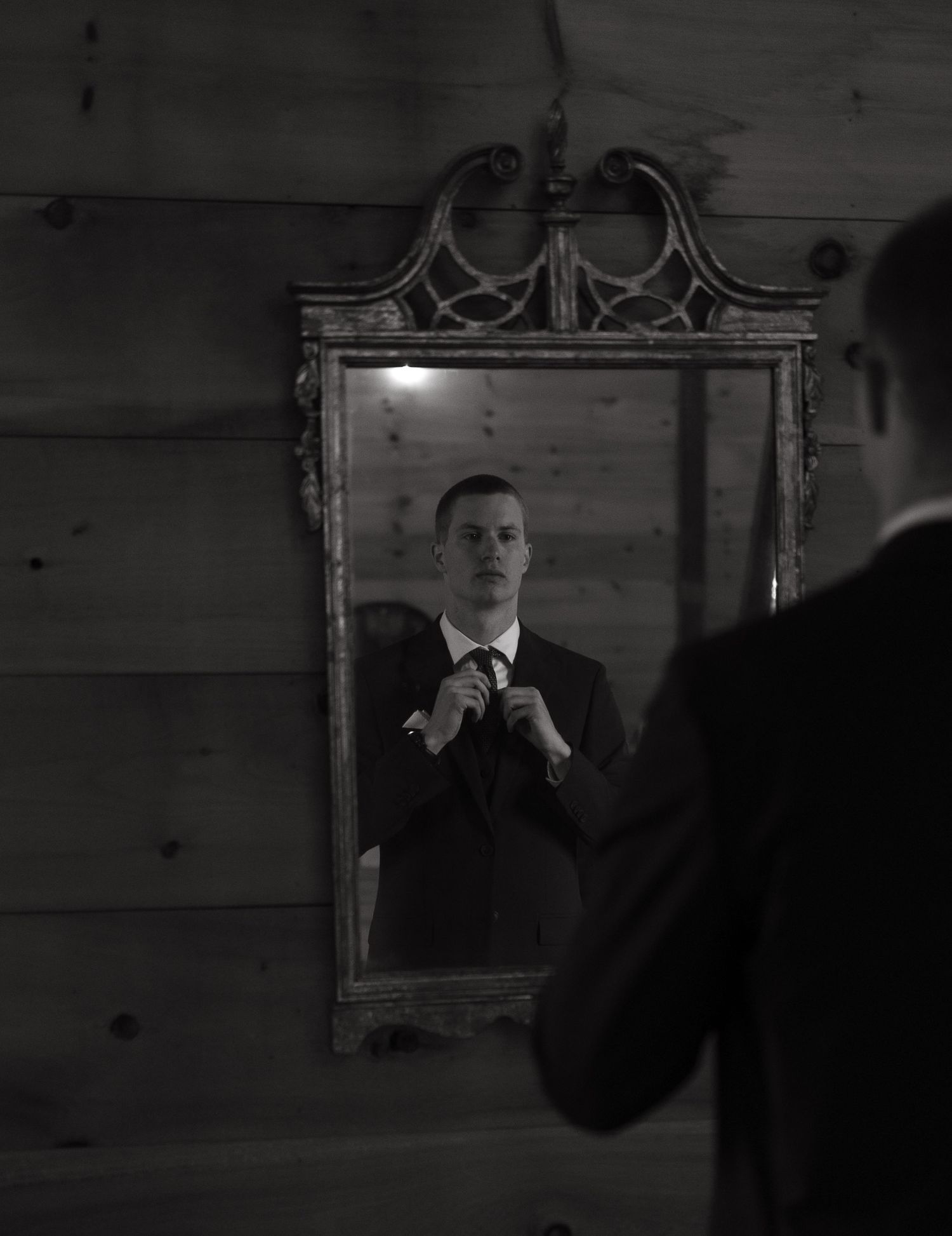 Black and white portrait of groom adjusting tie reflected in ornate vintage mirror on wood wall.