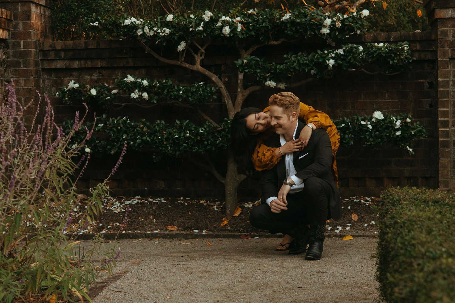 Couple sharing a joyful embrace in a garden with white flowering shrubs and brick wall backdrop.