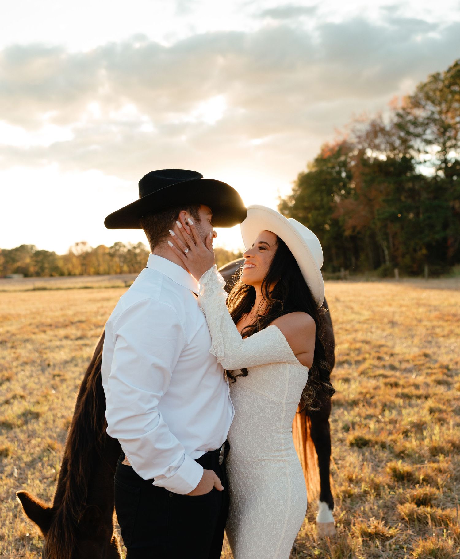 Smiling bride in white hat and dress embracing groom in black cowboy hat at golden sunset.