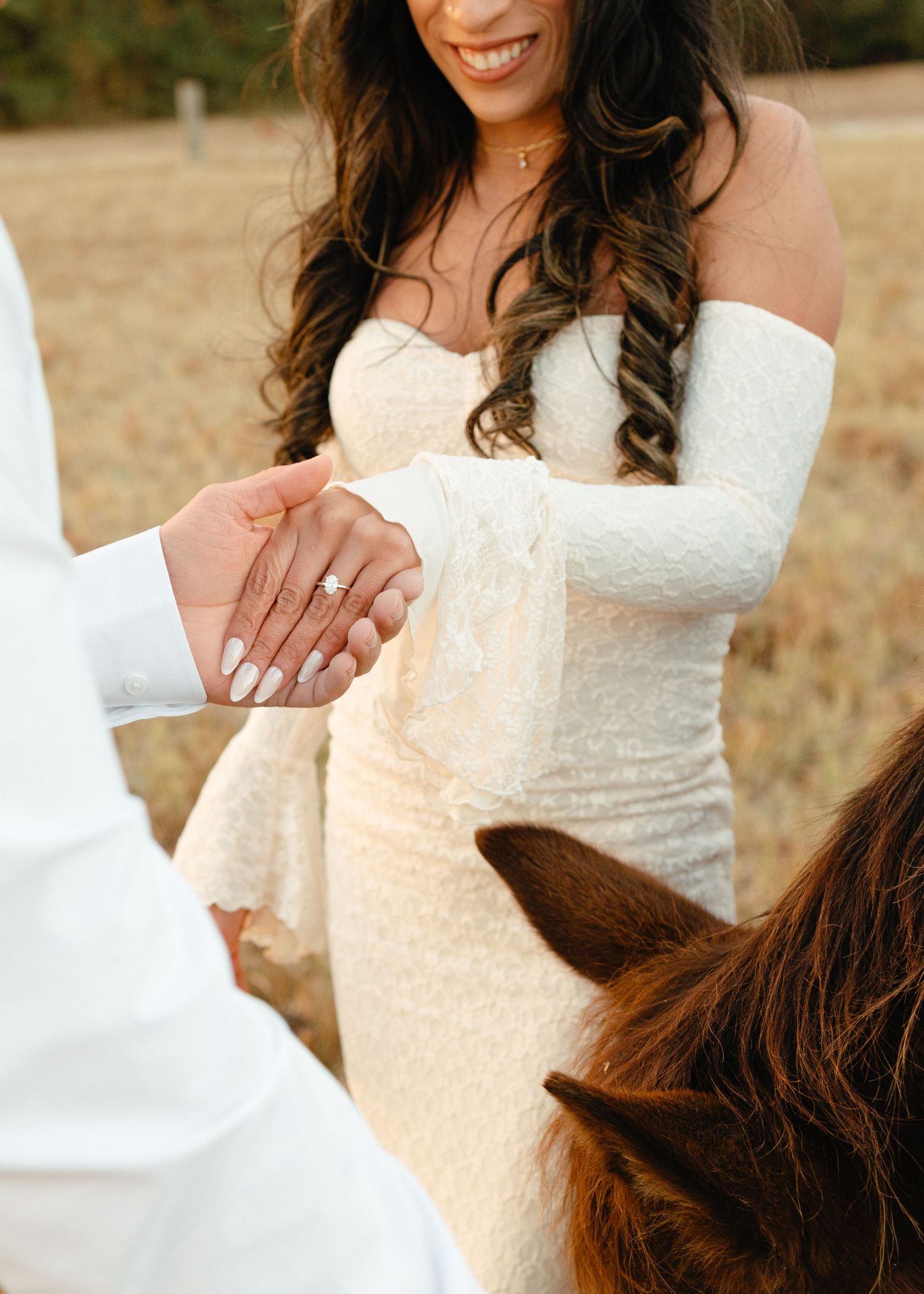 Close-up of bride and groom holding hands near a horse, highlighting her diamond engagement ring and lace wedding gown.