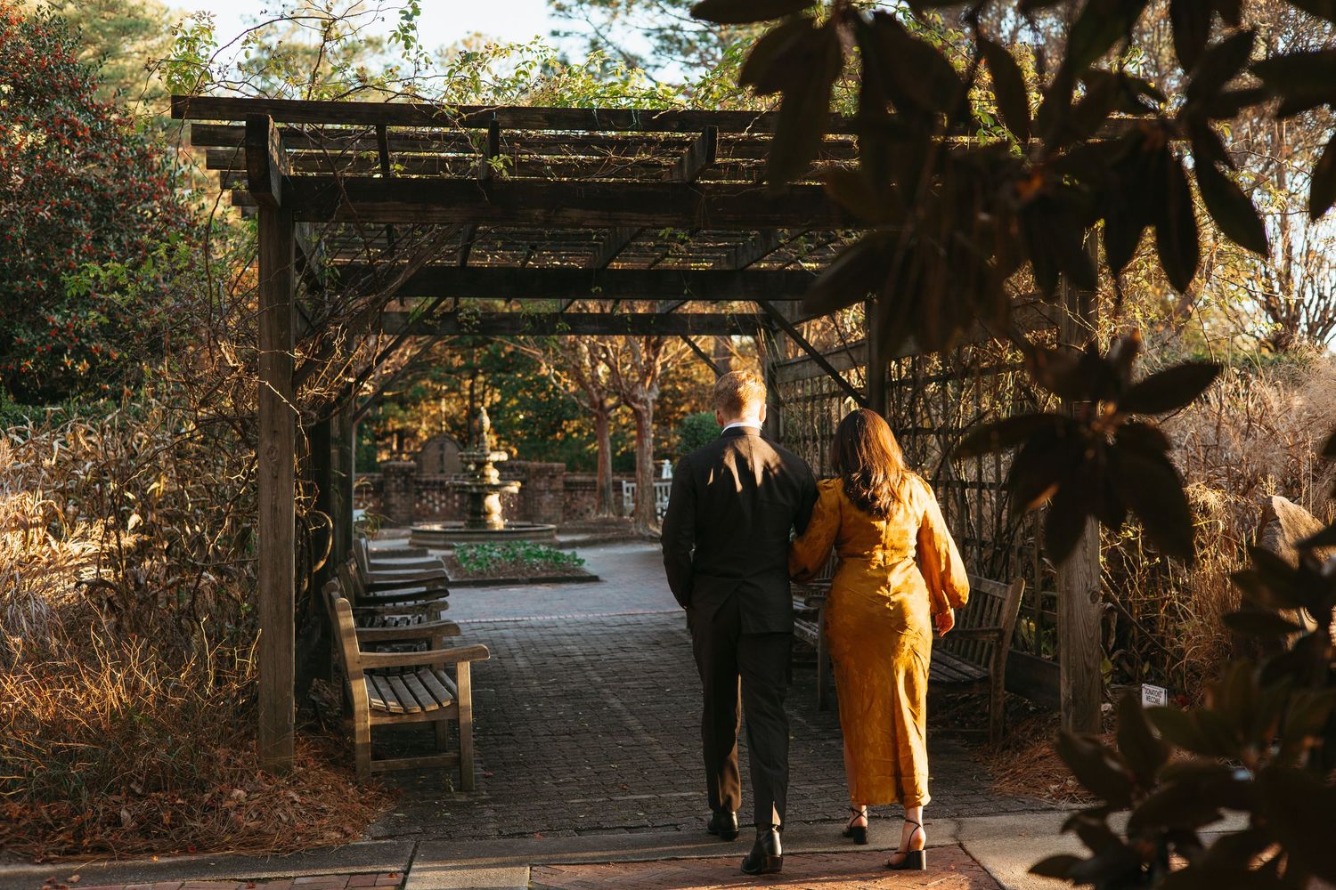 Two people walk hand-in-hand through a pergola-covered garden path surrounded by autumn foliage.