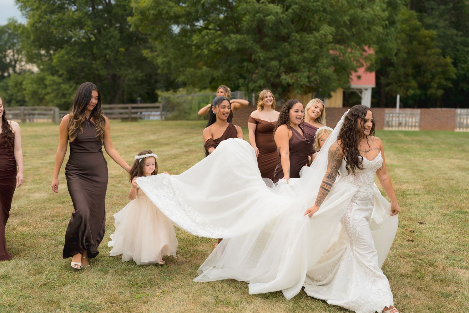 Bride in white gown walking joyfully with bridesmaids in brown dresses and flower girl on lawn.