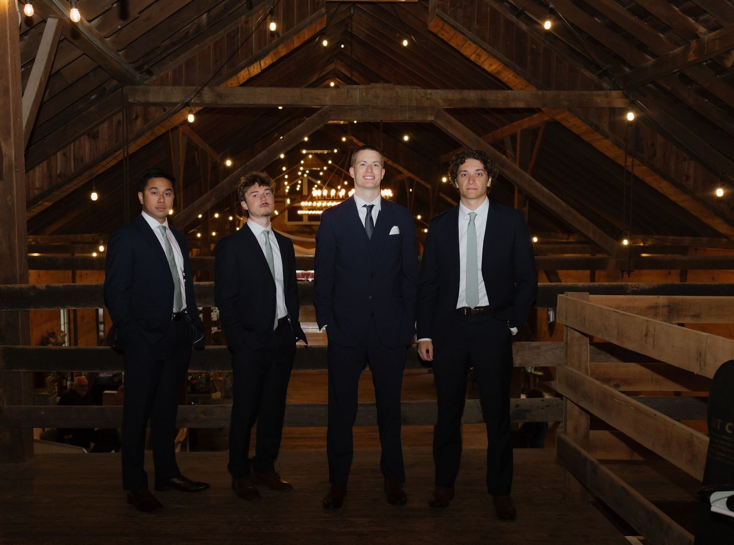 Groom and three groomsmen in dark suits pose inside rustic wooden barn with string lights overhead.