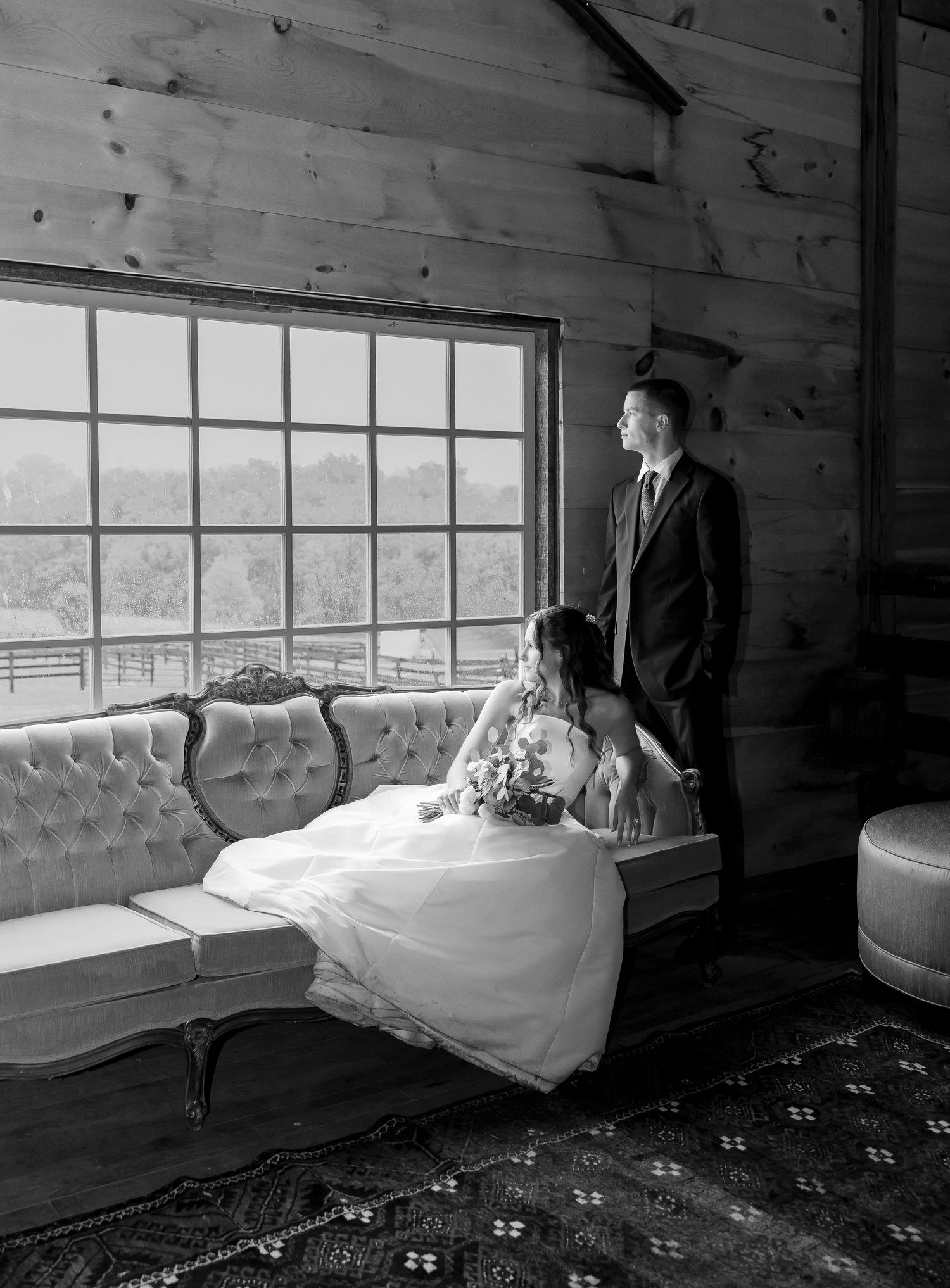 Black and white portrait of bride and groom gazing out a large window in a rustic barn.