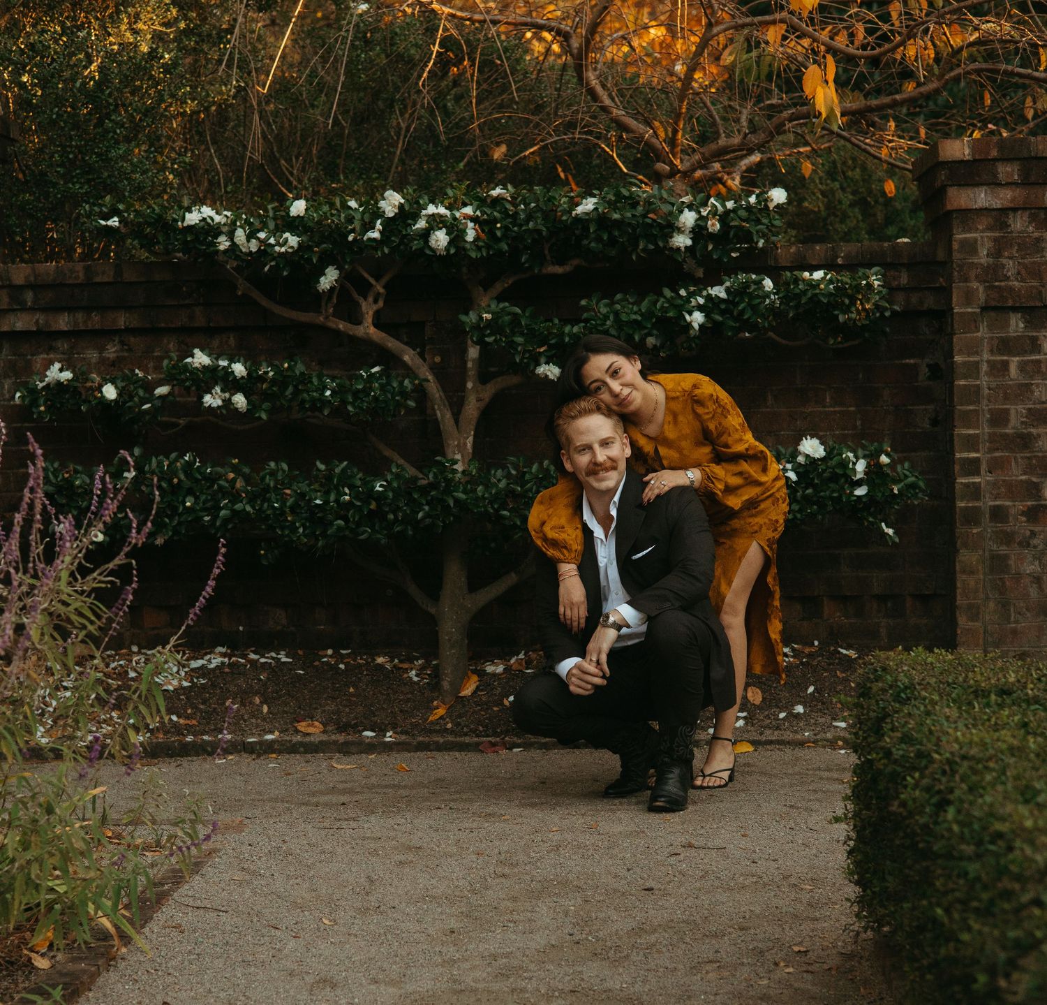 Couple posing playfully in front of a white flowering wall in a lush garden at dusk.