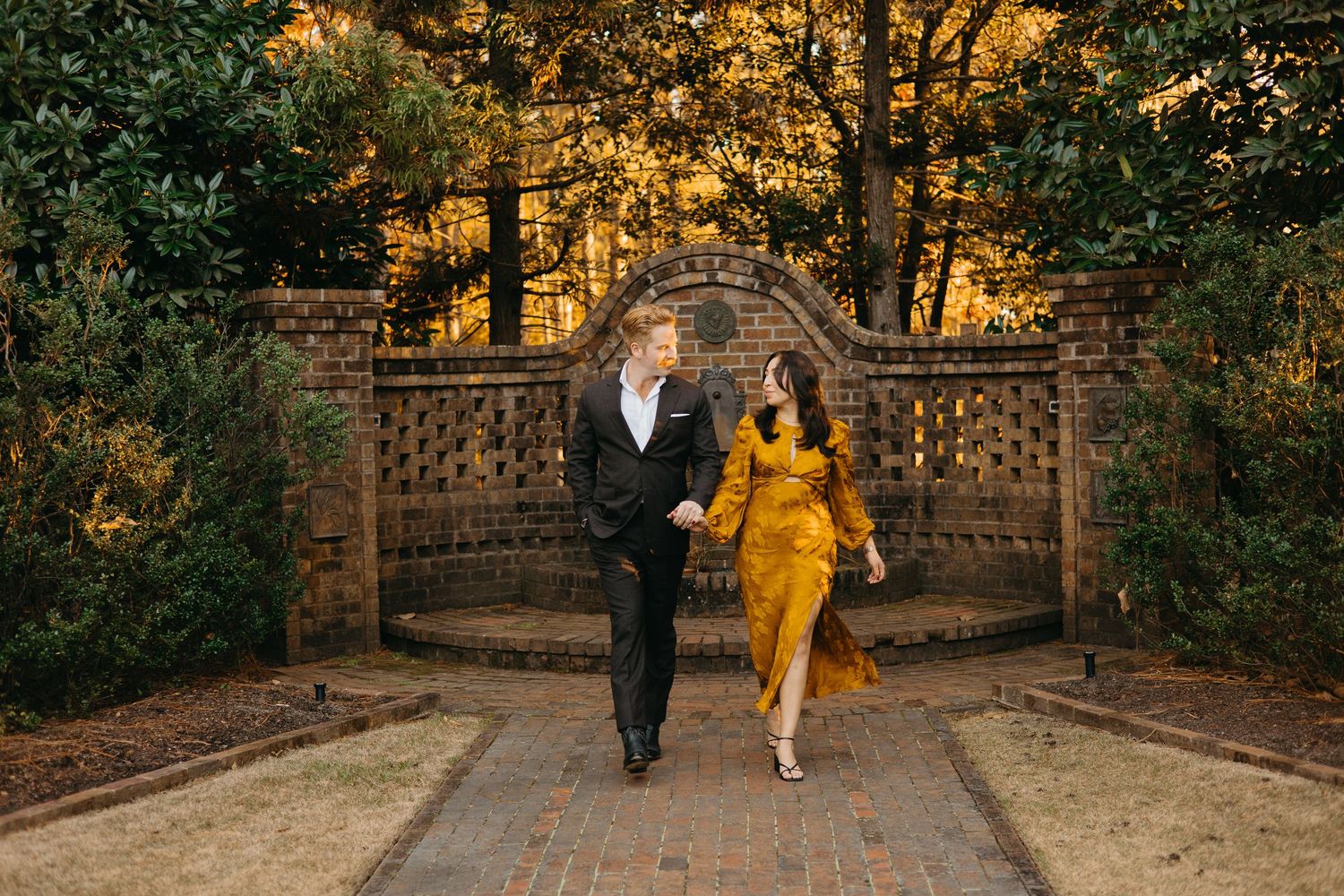 Couple laughing and walking together on brick path through garden with golden autumn trees.
