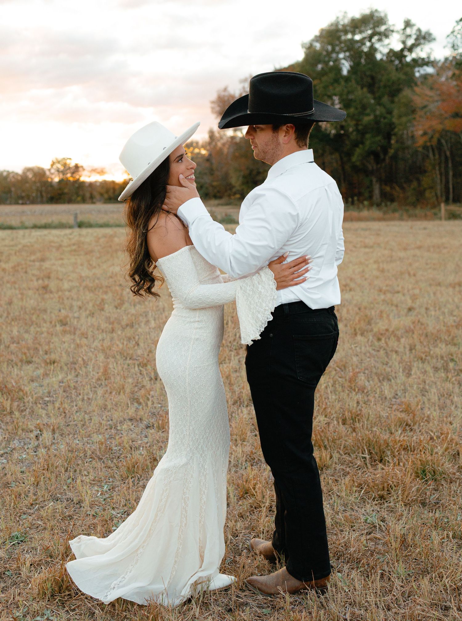 Bride in white gown and wide-brim hat smiles at groom in black cowboy hat during sunset field photoshoot.