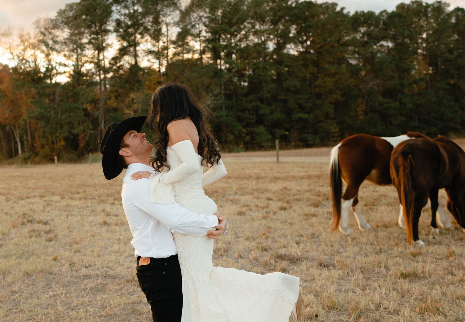 Groom lifting bride in white dress while a horse grazes in the golden field behind them.