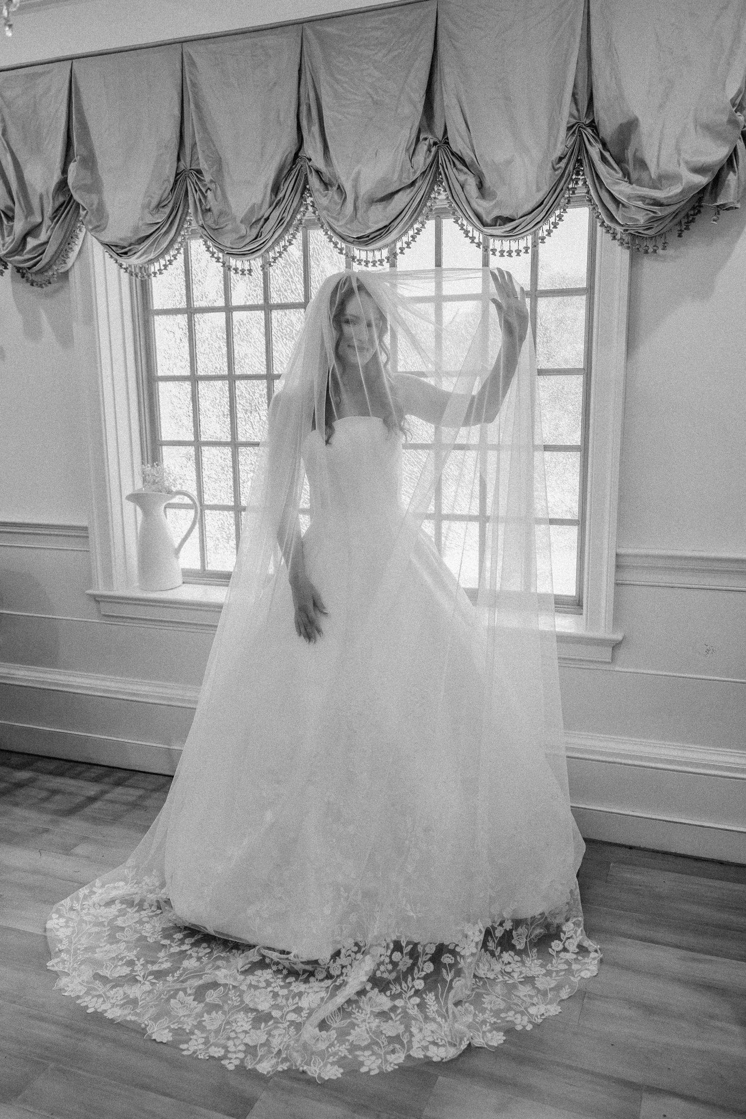 Elegant bride in lace ball gown lifting veil in black and white portrait near draped windows.