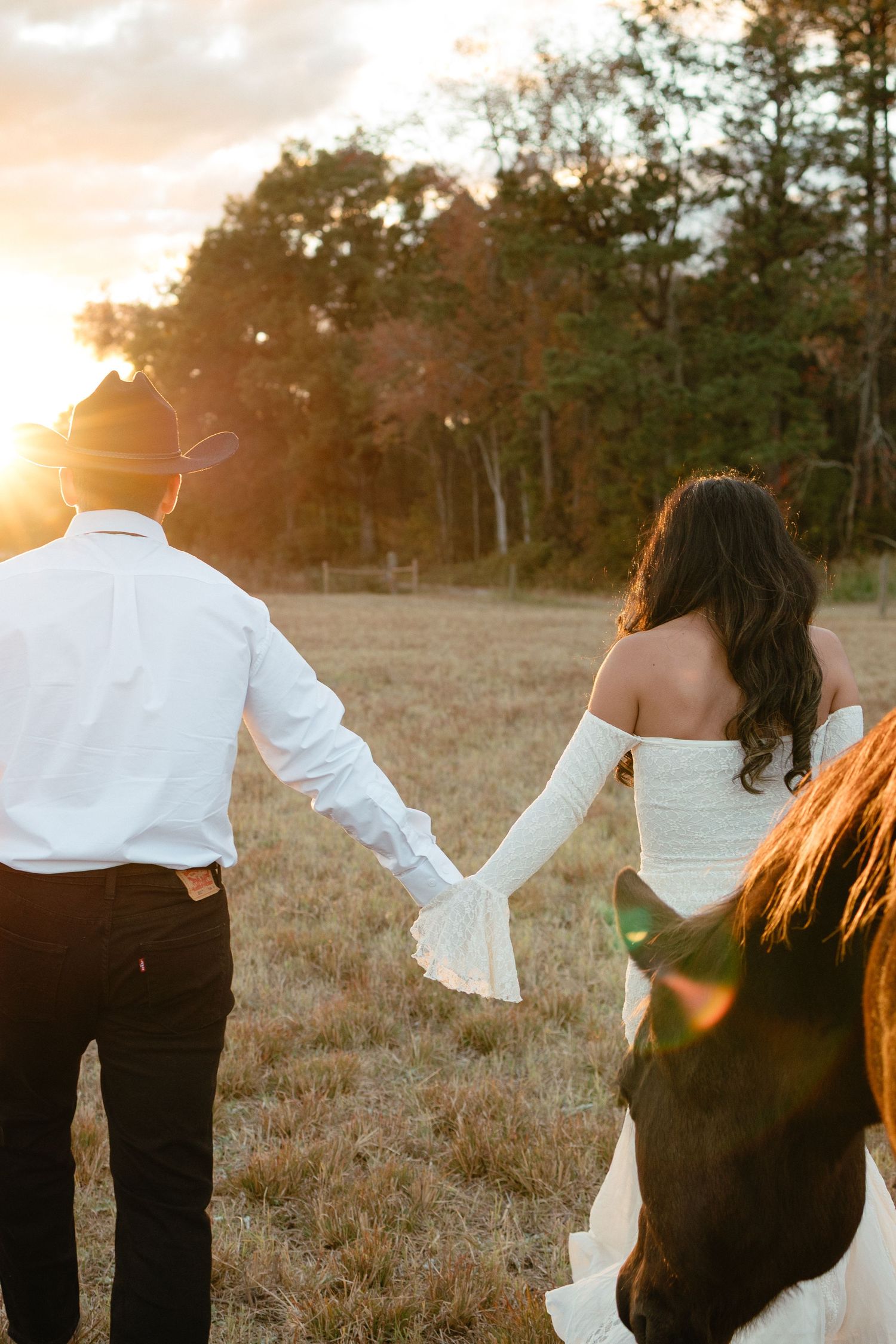 Couple holding hands walking toward a glowing sunset in a field with a horse nearby.