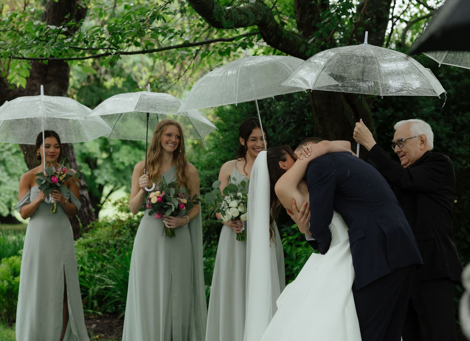 Bridesmaids in sage green gowns hold clear umbrellas as bride and groom share a kiss in the rain.