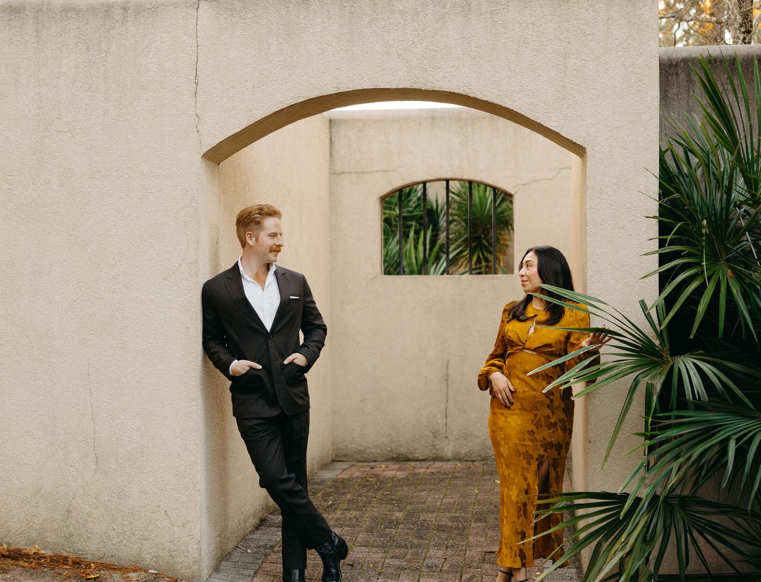 Couple laughing together near a stucco archway with tropical palm plants during an outdoor photo session.
