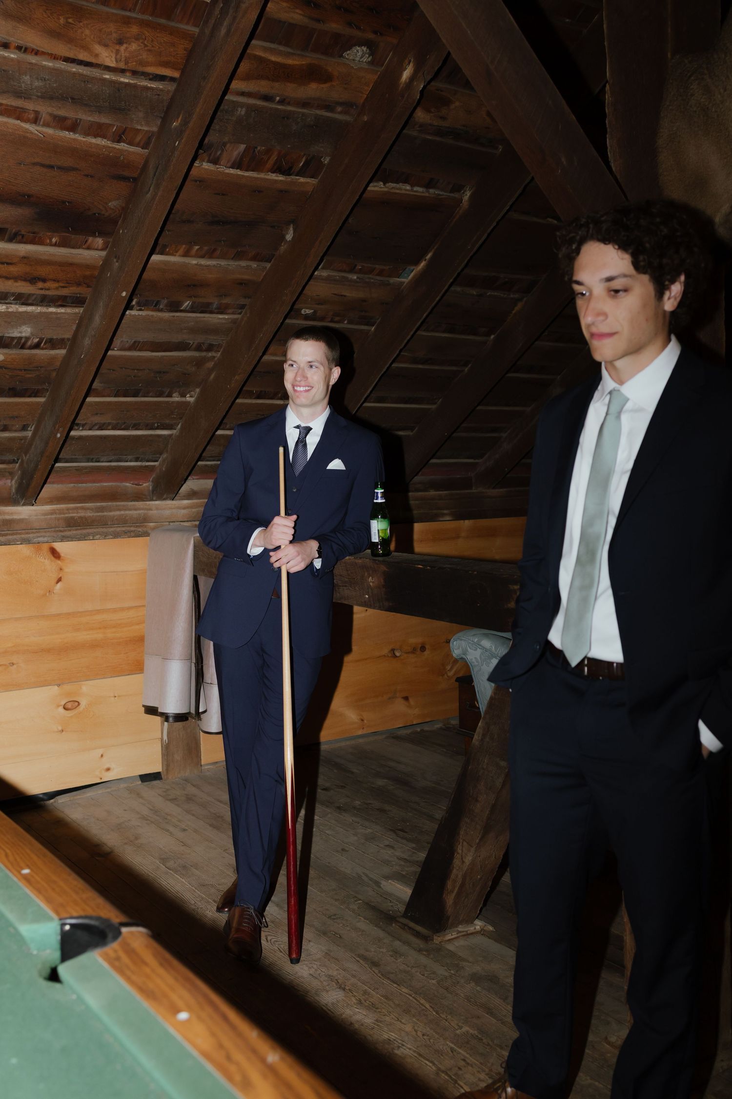 Two groomsmen in suits playing pool in a rustic wooden barn loft space.