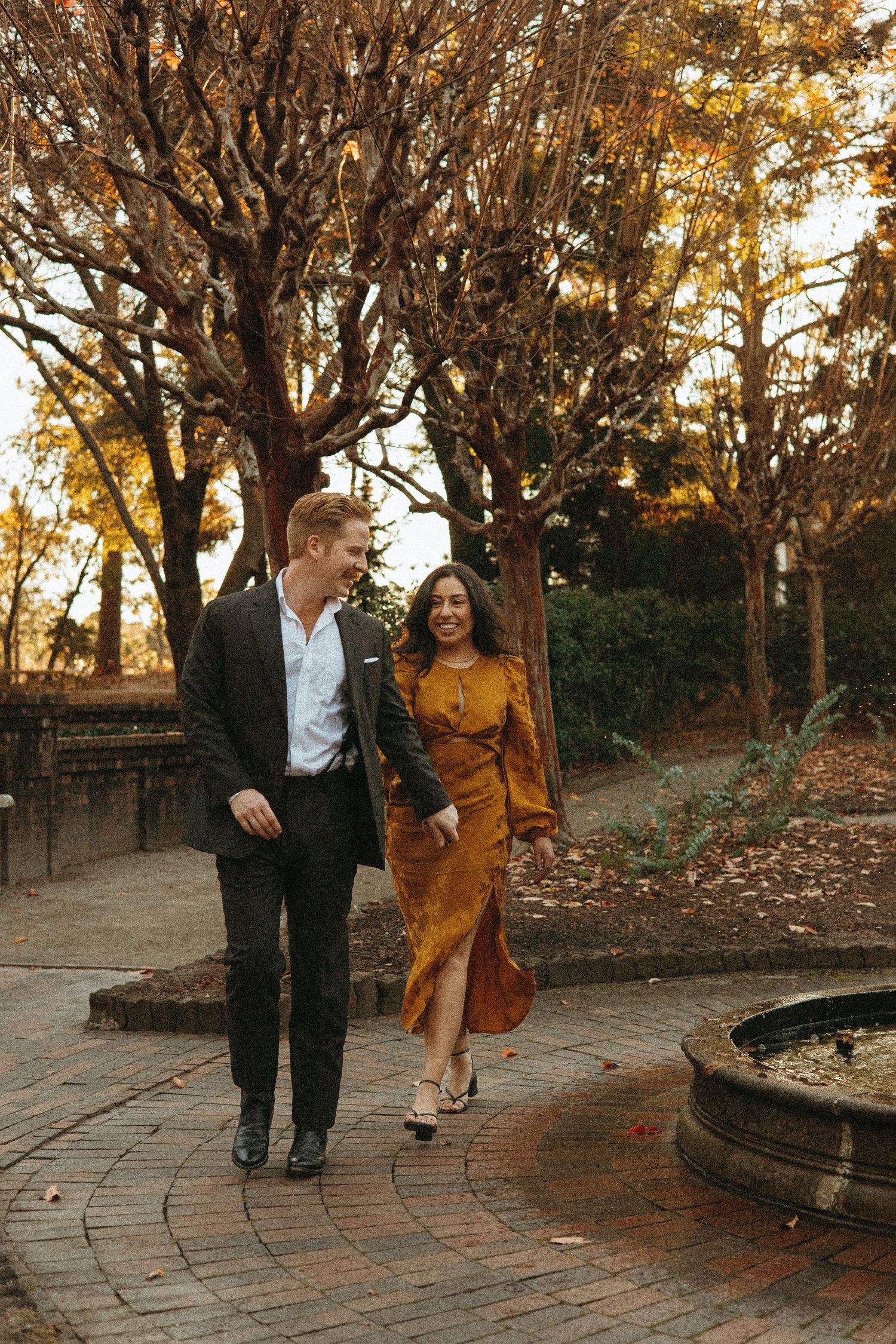 Smiling couple strolling through a sunlit autumn garden, woman in golden dress, man in dark suit.