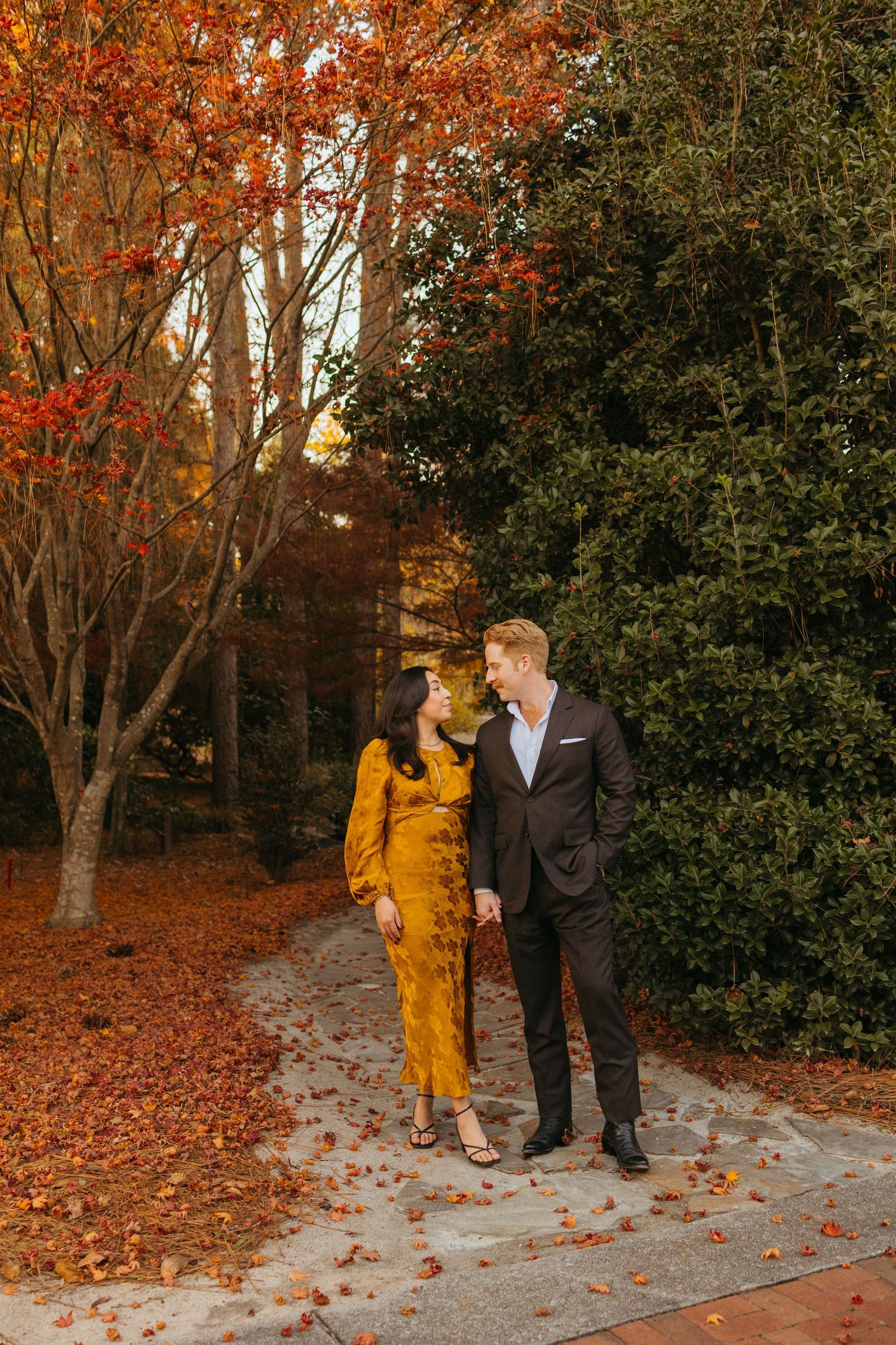 Couple holding hands and gazing at each other on a path lined with brilliant red autumn trees and evergreens.