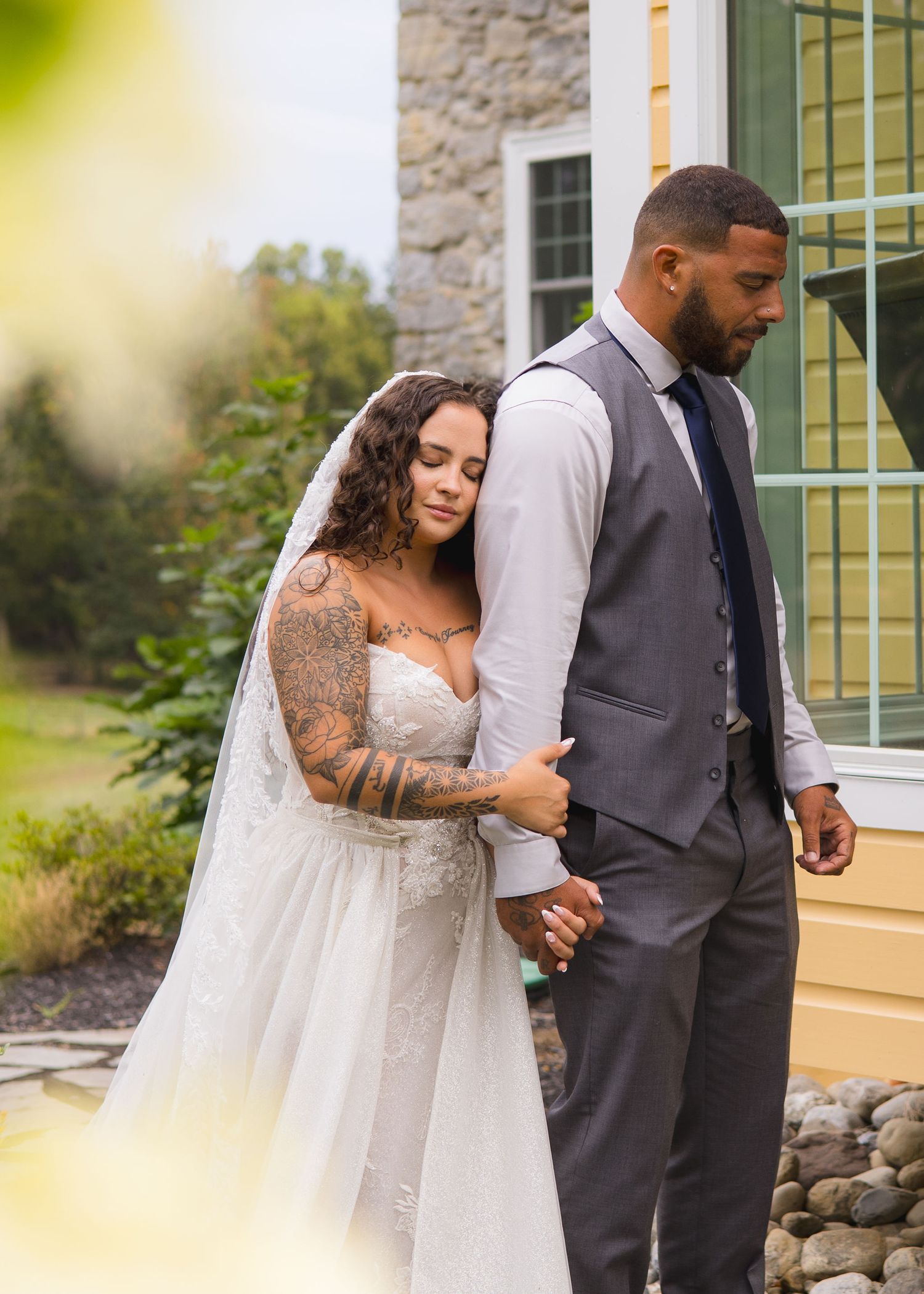 Tattooed bride in lace gown embracing groom in gray vest while holding hands outdoors at stone venue.