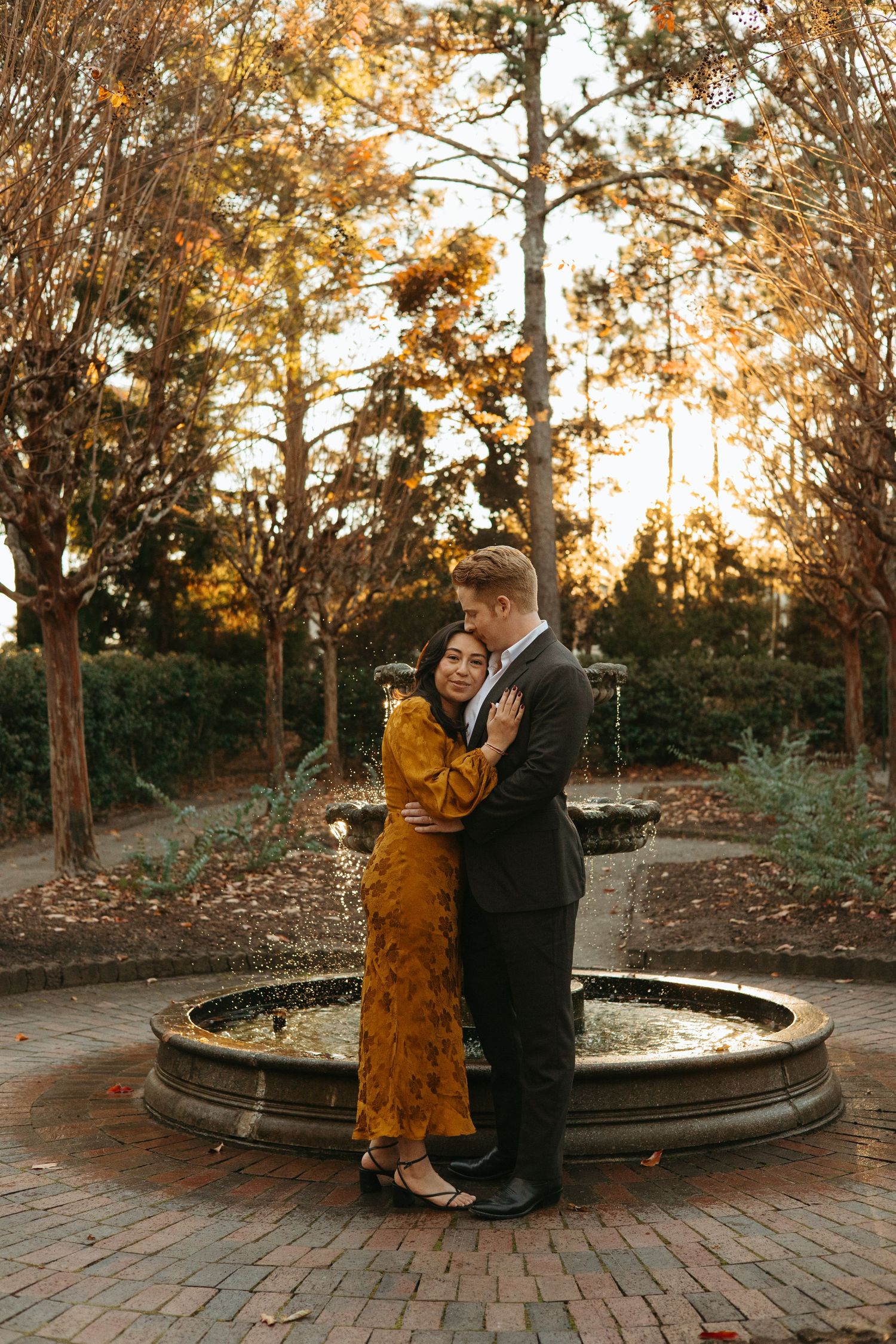 Couple embracing by a stone fountain in an autumn garden, woman in golden dress, man in dark suit.