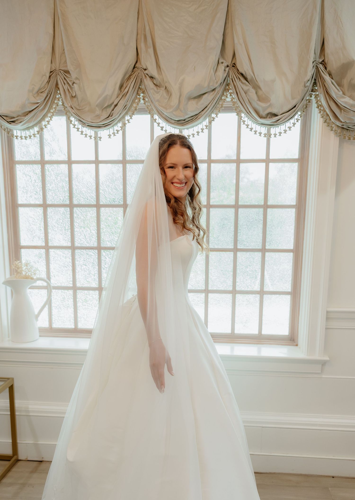 Radiant bride in white ballgown and cathedral veil smiling before large elegant window.