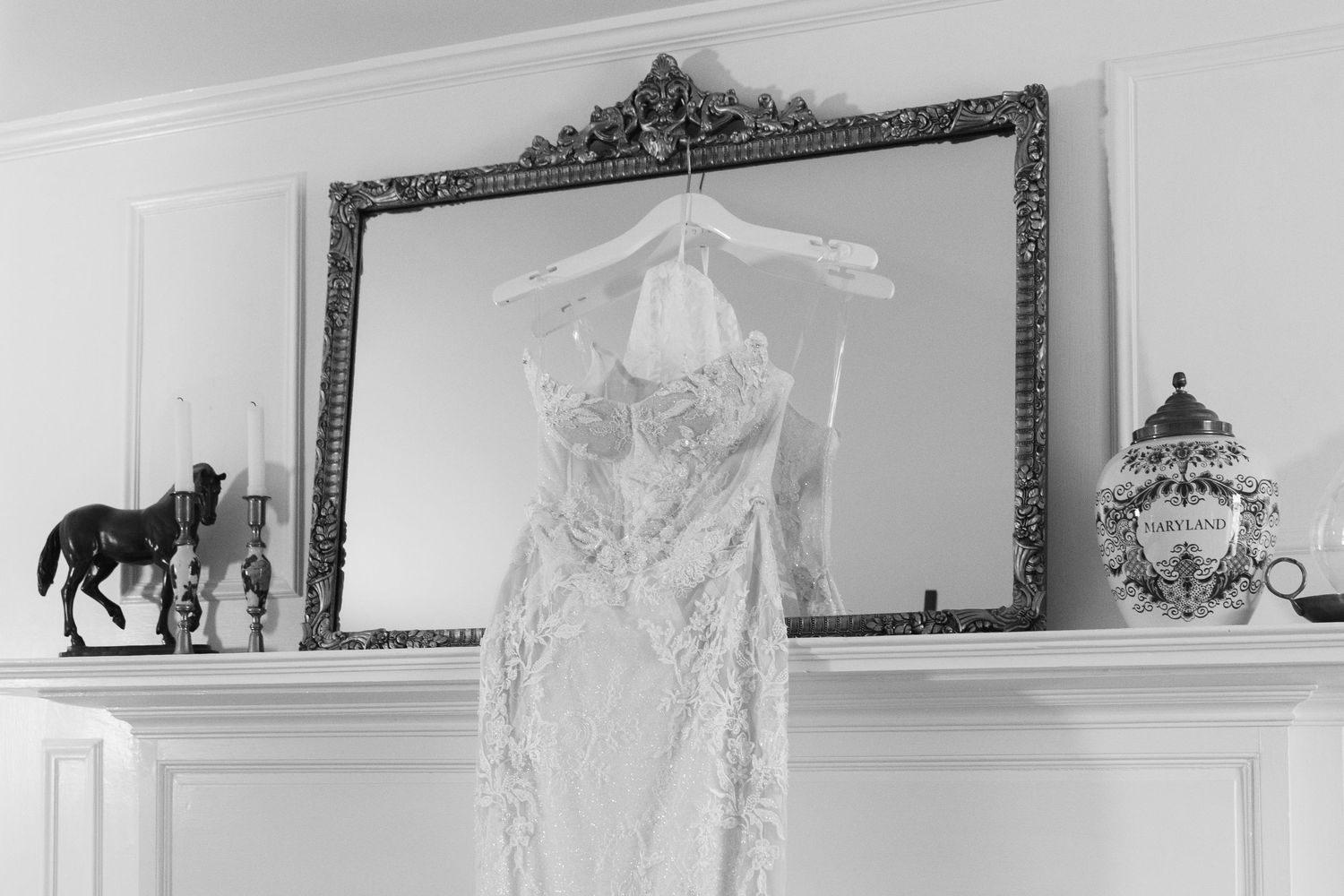 Black and white photo of an elegant lace wedding dress hanging on a ornate mirror above a fireplace mantel.