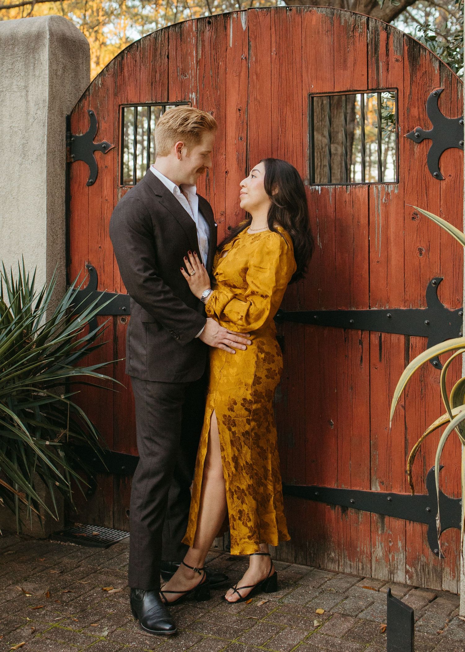Couple embracing before a rustic red wooden gate, man in dark suit and woman in golden yellow floral dress.