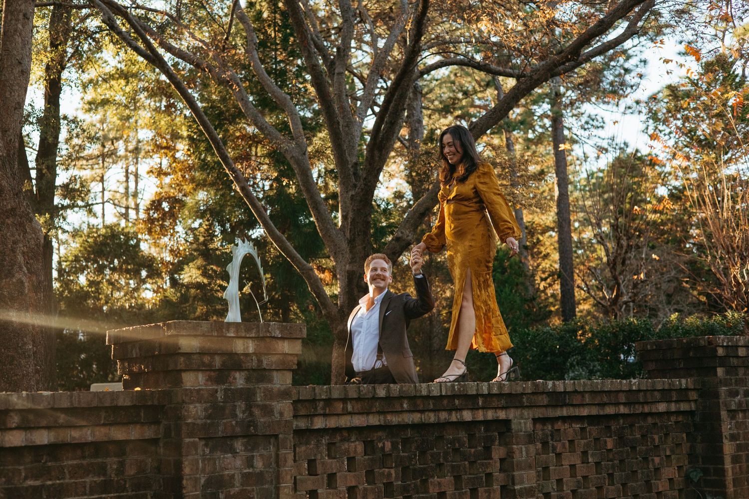 Playful couple posing on a brick garden wall surrounded by tall autumn trees.