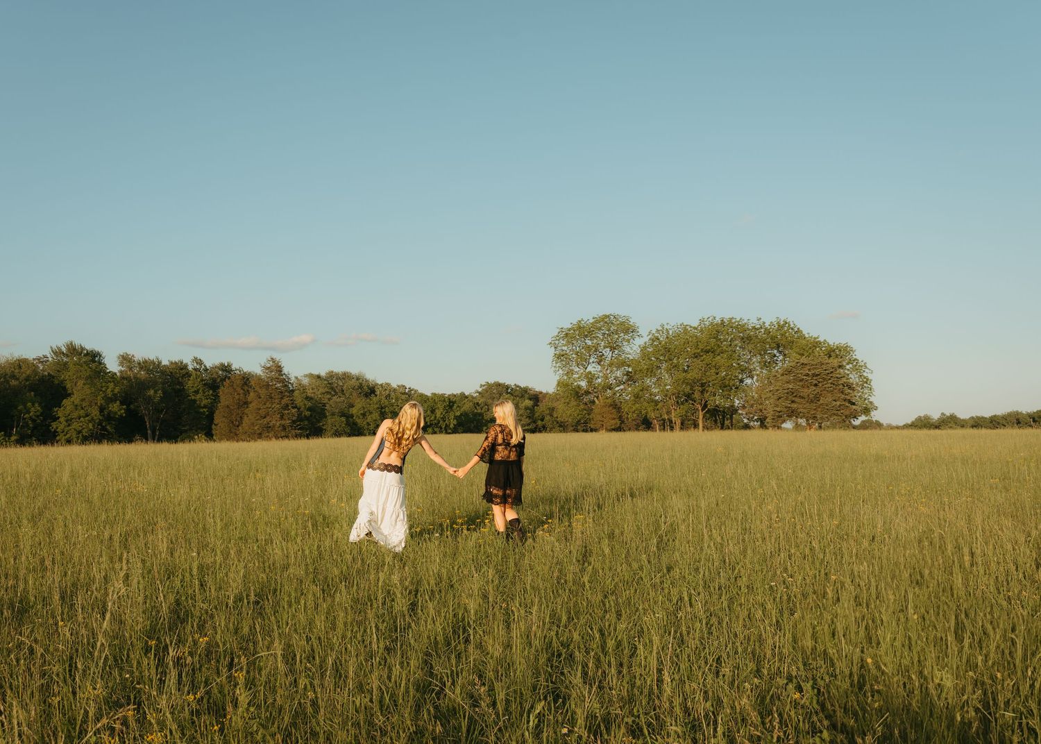 Two young women holding hands walking through a lush green field with trees in the background on a sunny day.