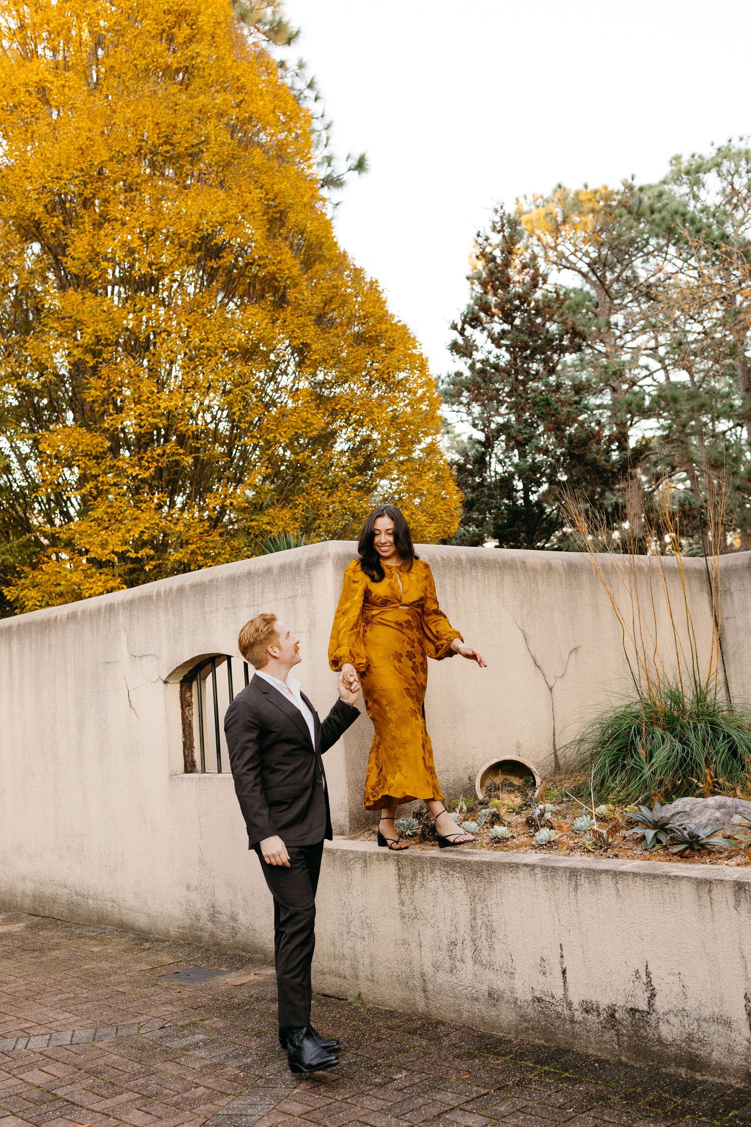 Man in black suit holds woman's hand as she stands on a wall in a gold dress amid vibrant autumn foliage.