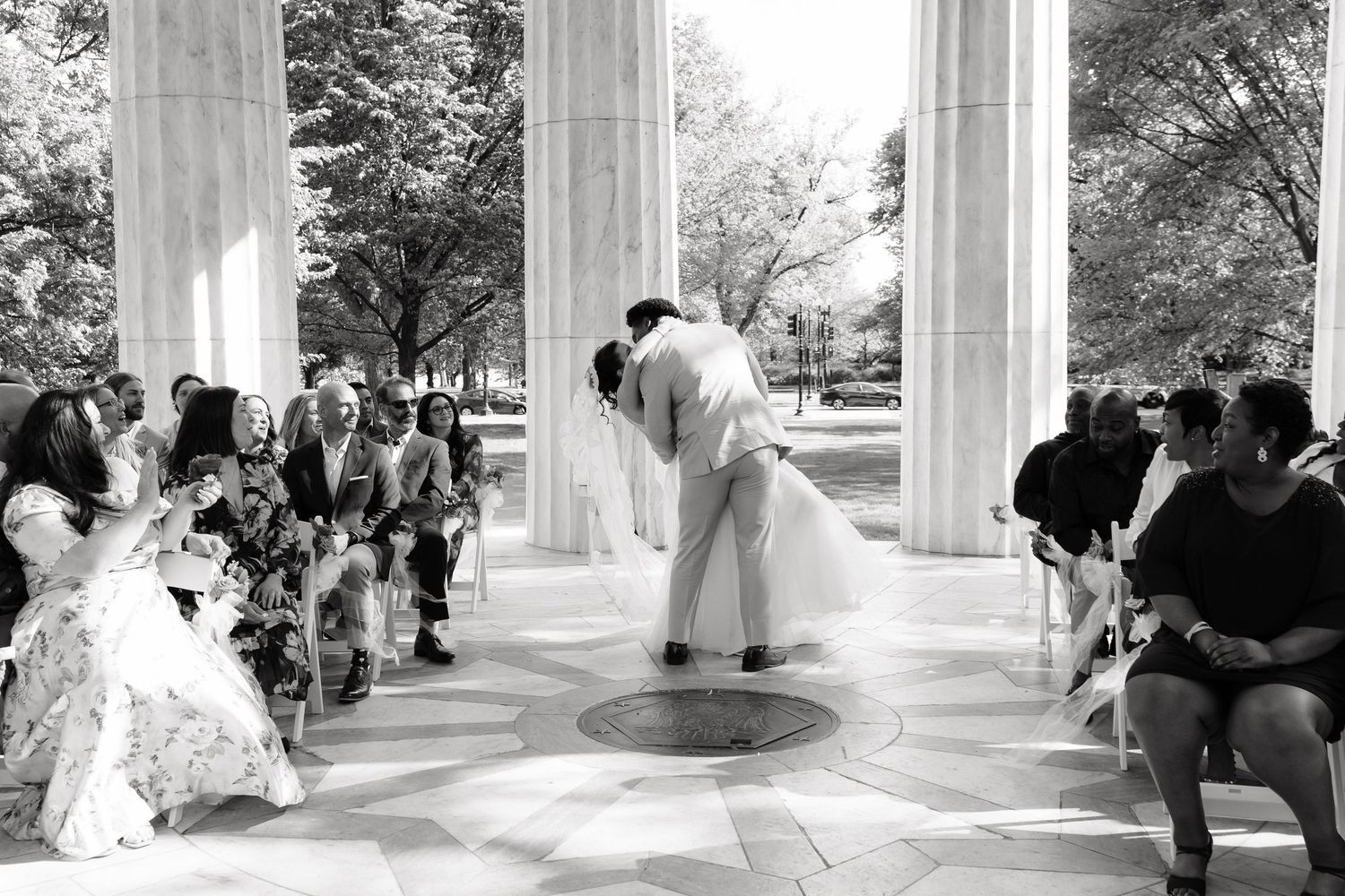Black and white photo of bride and groom sharing first kiss at outdoor columned wedding ceremony.