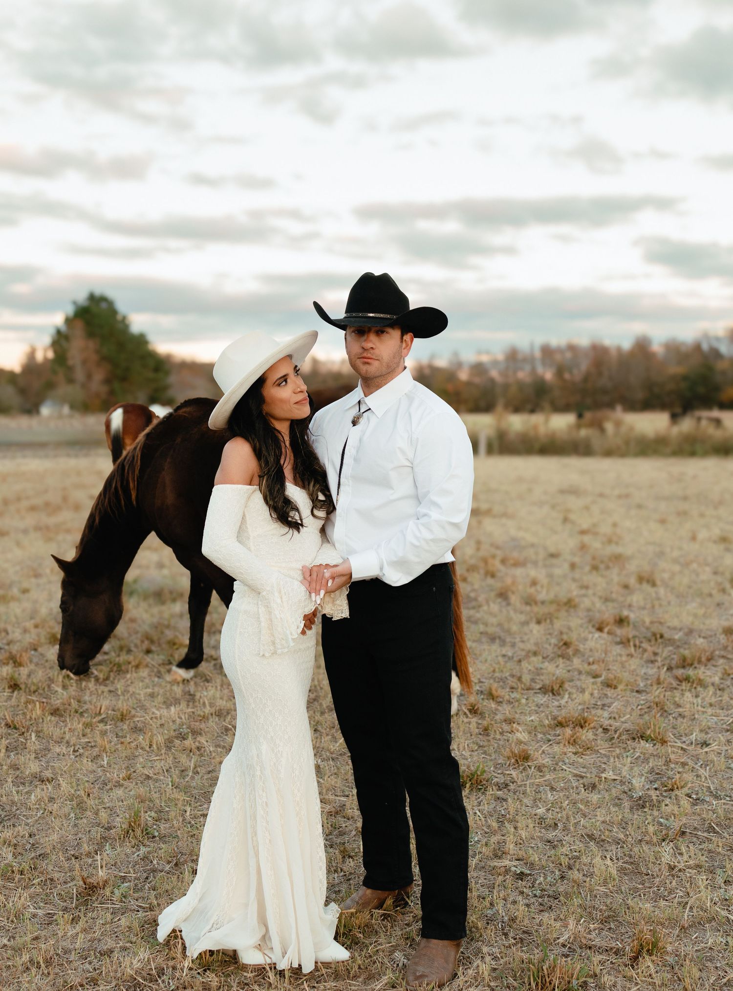 Western wedding couple standing together in field with brown horse grazing behind them.