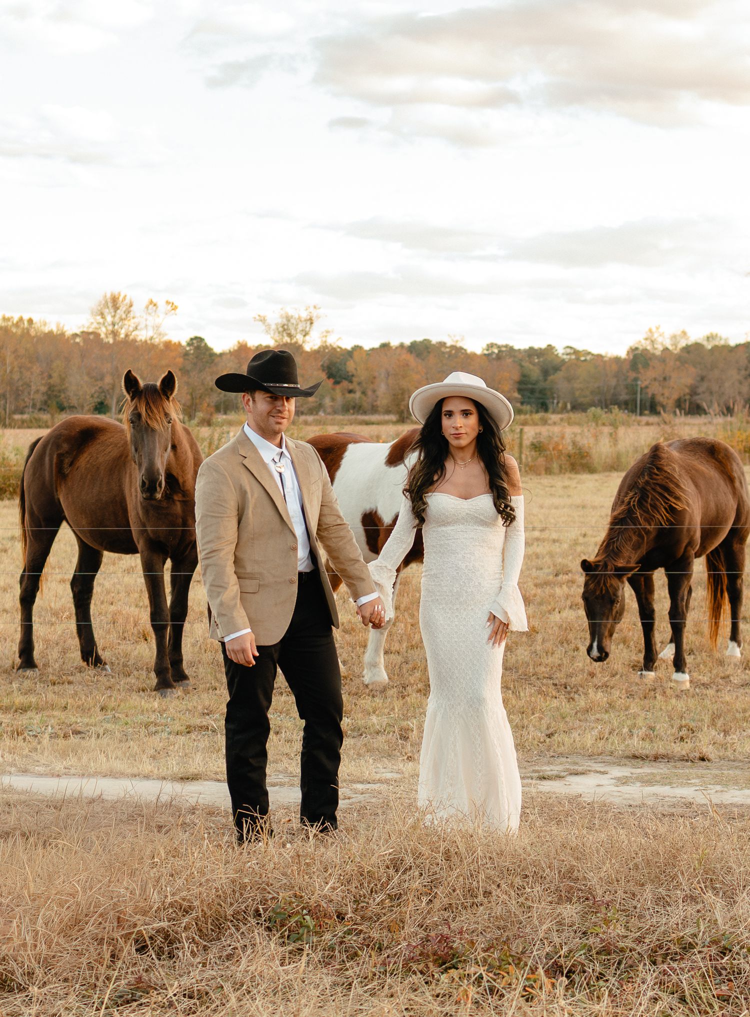 Bride and groom in cowboy hats holding hands in a field with brown horses grazing behind them at golden hour.