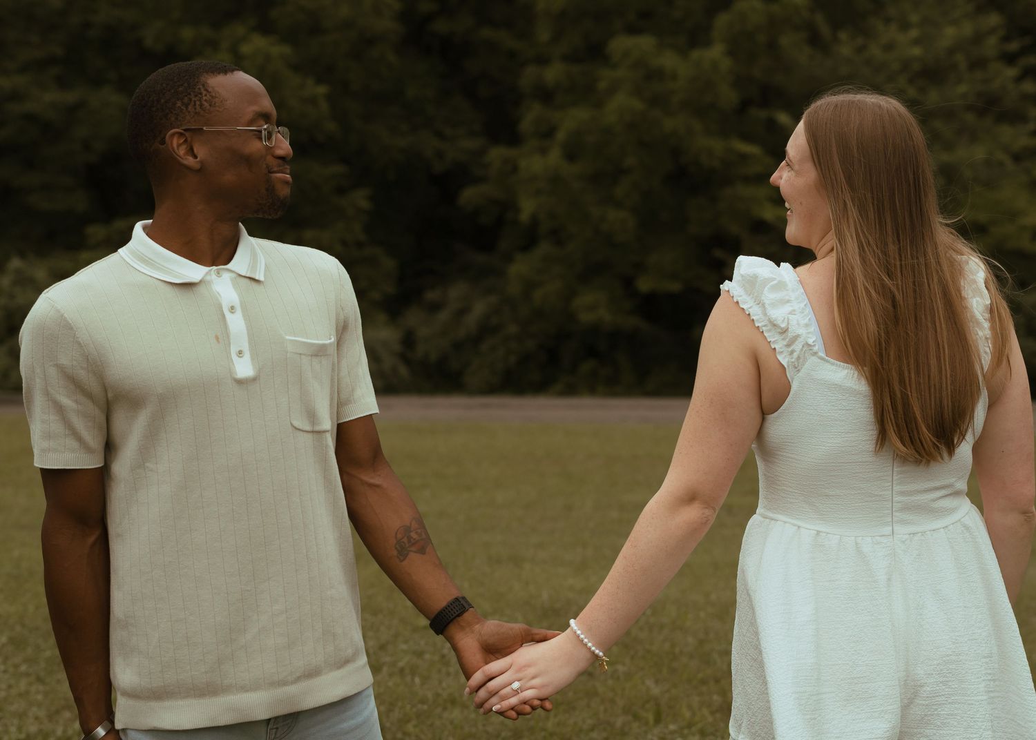 Interracial couple holding hands and smiling at each other outdoors in a green park setting.