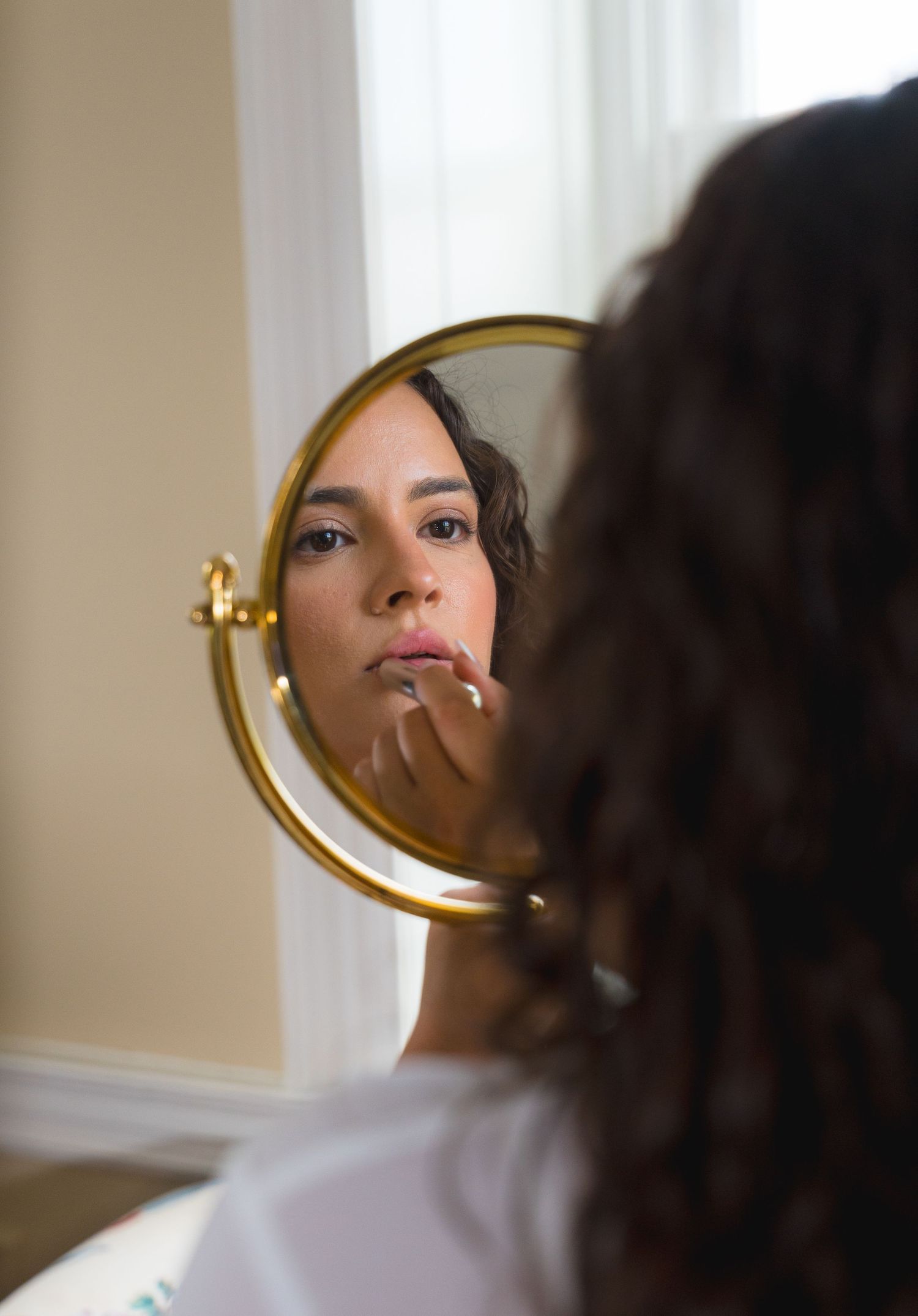 Young woman applying lip makeup while looking into a round gold-framed vanity mirror.