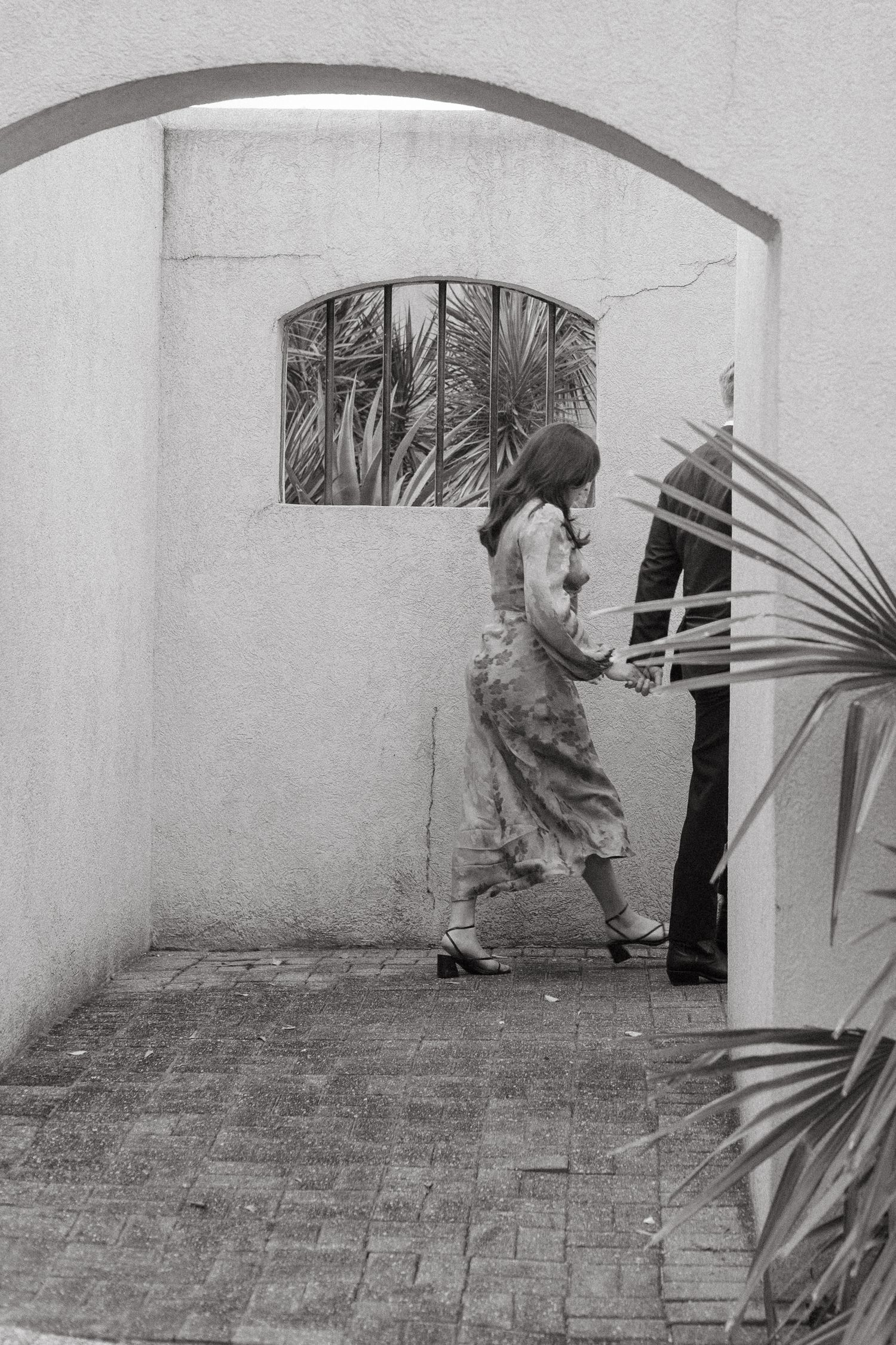 Black and white photo of a couple holding hands while walking through a stucco archway with palms.