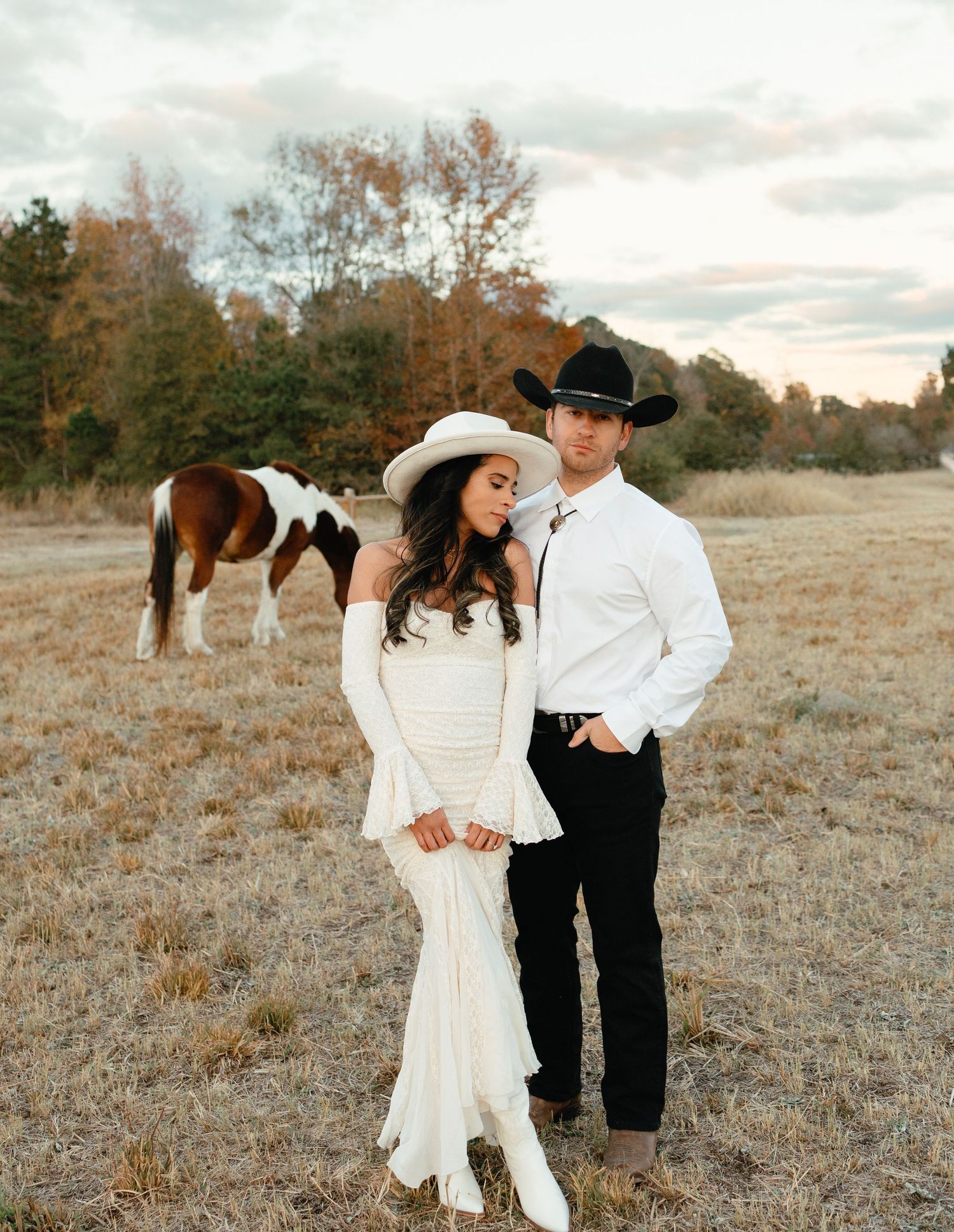 Bride and groom posing in western attire with paint horse grazing behind them at sunset.