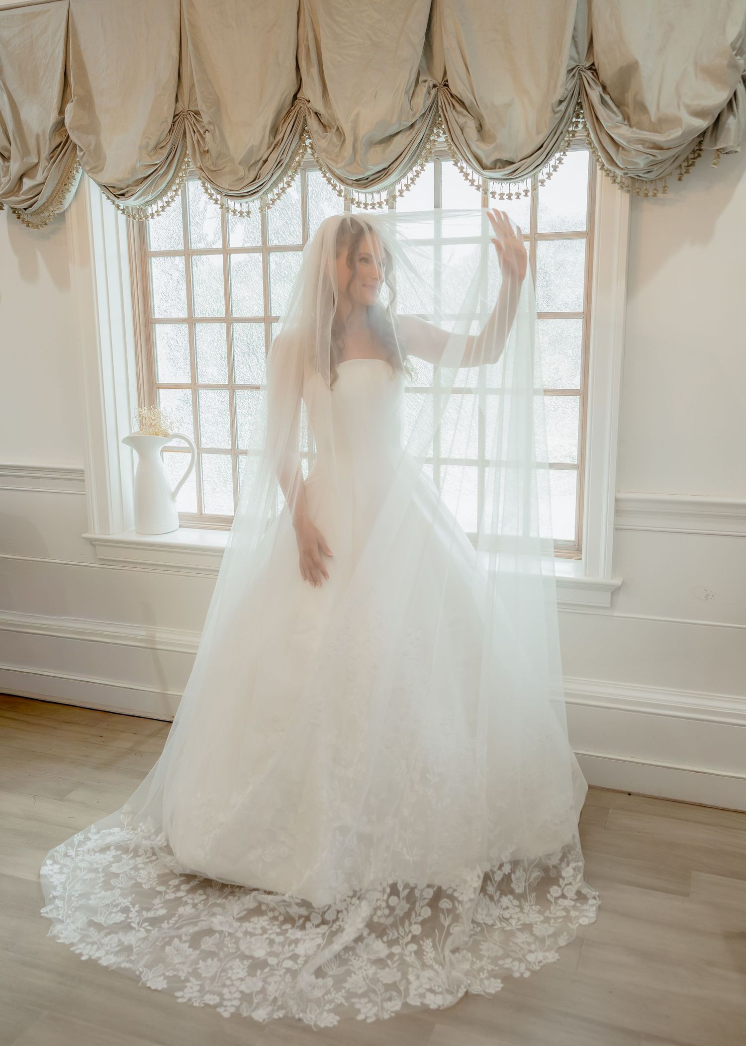 Elegant bride in lace ball gown and cathedral veil poses by large window in softly lit room.