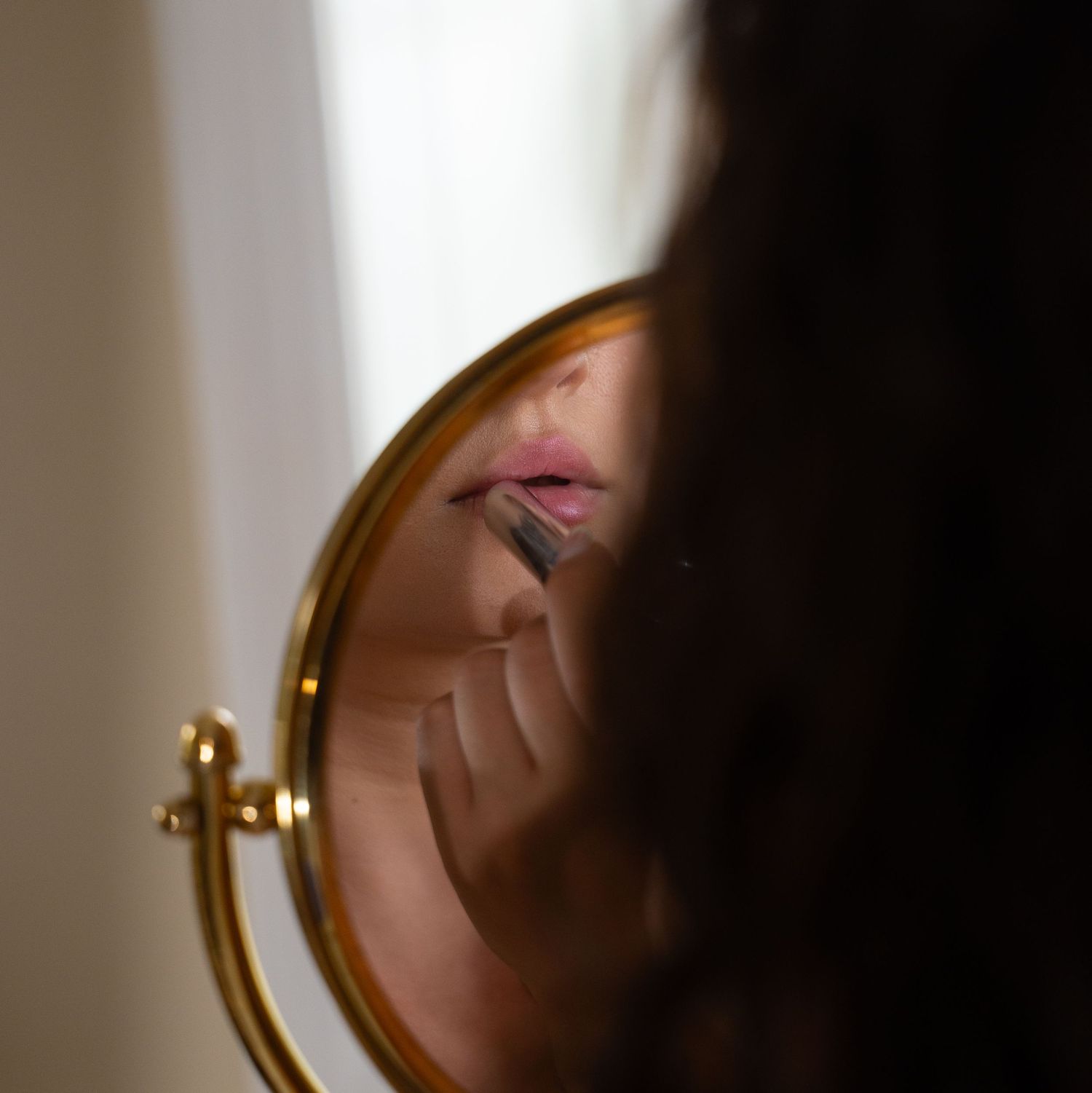 A woman applies lipstick while looking at her reflection in a round gold-framed vanity mirror.
