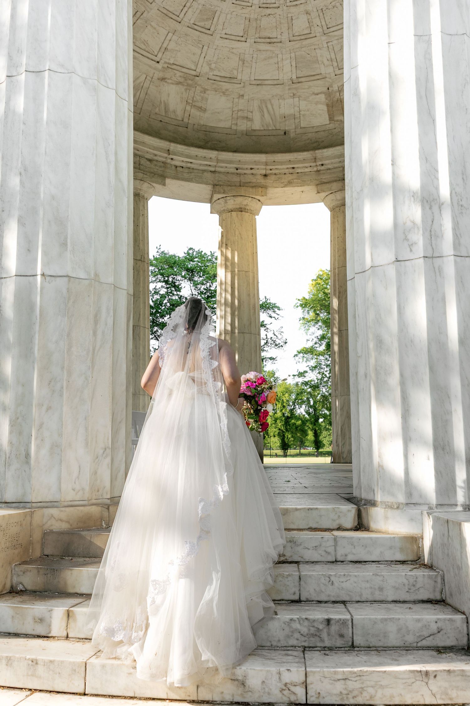 Bride in white gown and veil holding bouquet standing on marble steps of a classical rotunda.