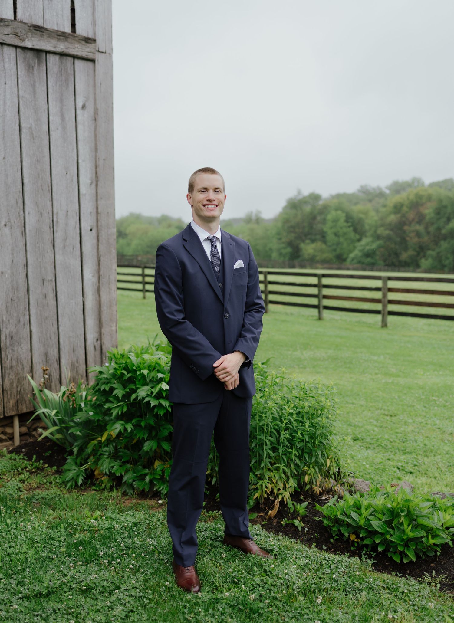 Dapper groom in navy three-piece suit standing on lush green lawn beside rustic barn.