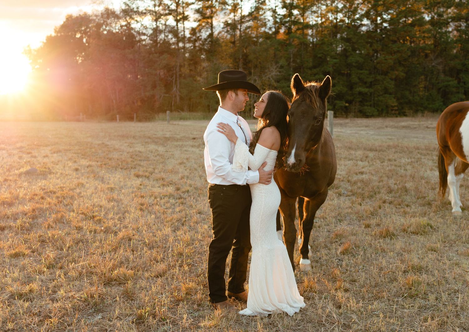 Couple in western wedding attire sharing a romantic moment beside a brown horse at sunset.
