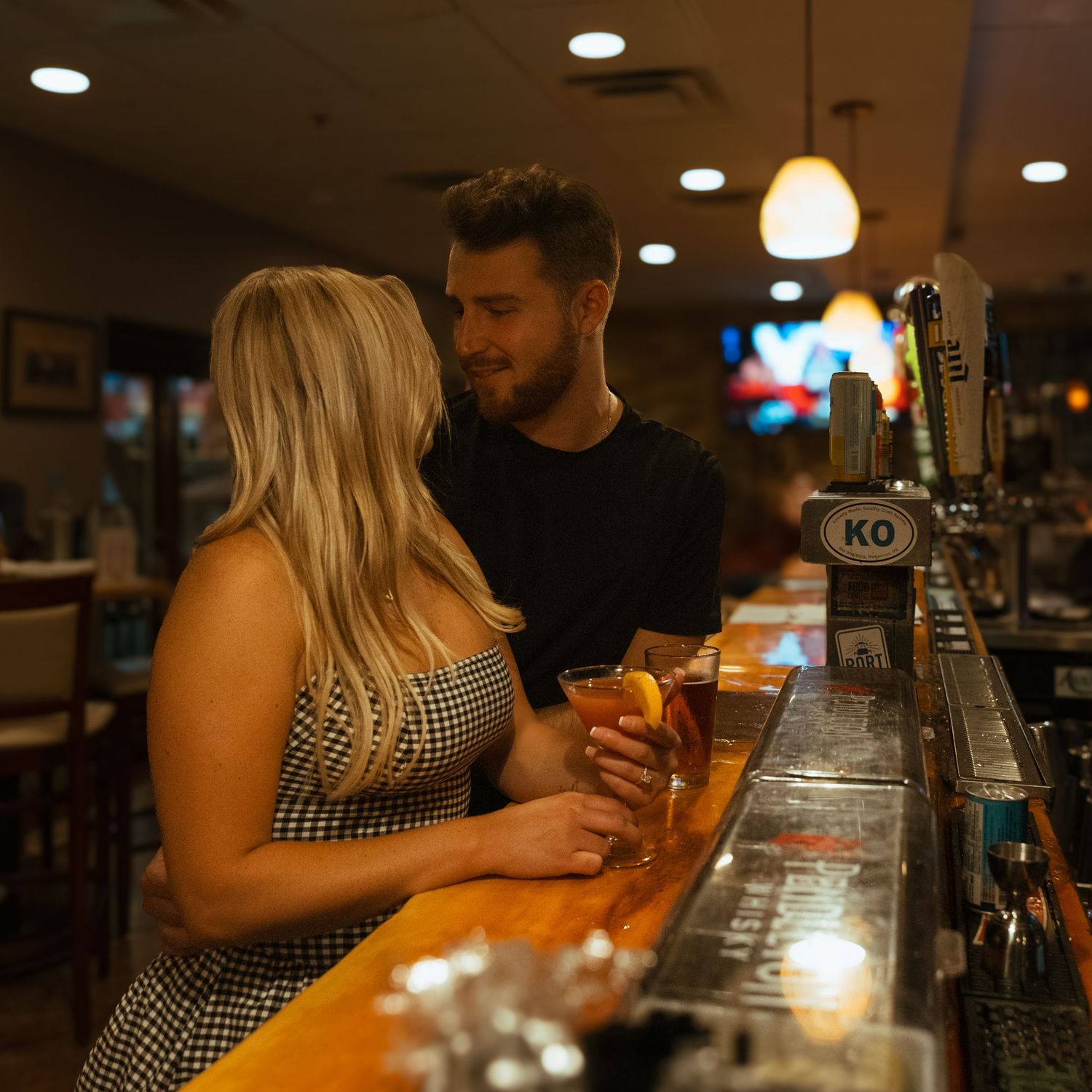 Couple enjoying drinks together at a warmly lit bar counter with beer taps and cocktails in hand.
