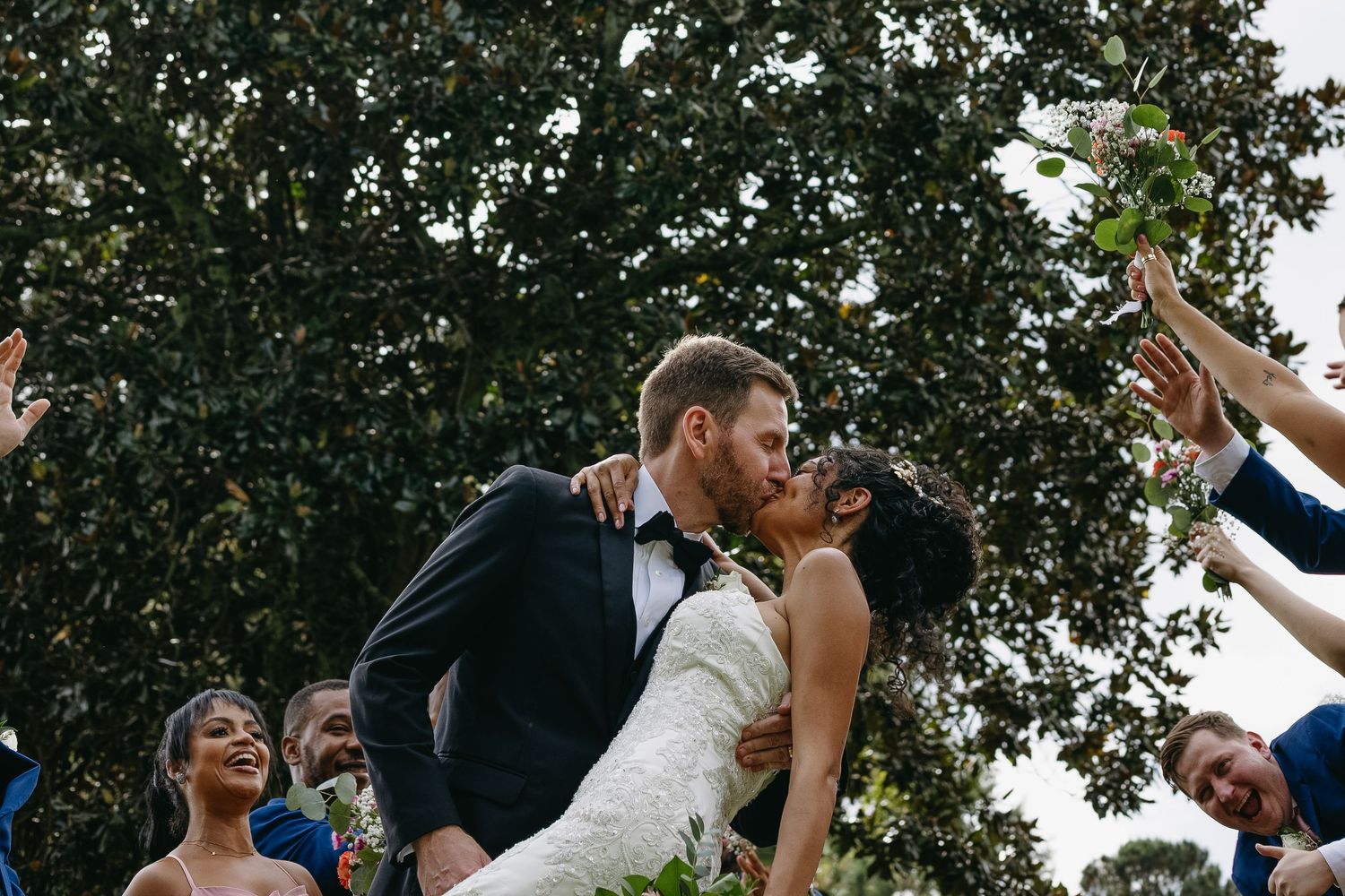 Bride and groom share a romantic kiss surrounded by cheering wedding guests outdoors under lush green trees.