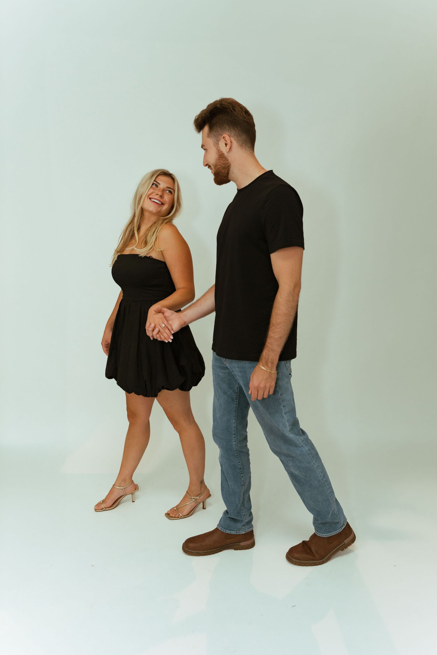Couple holding hands in matching black outfits, woman in mini dress and man in jeans, smiling against white background.