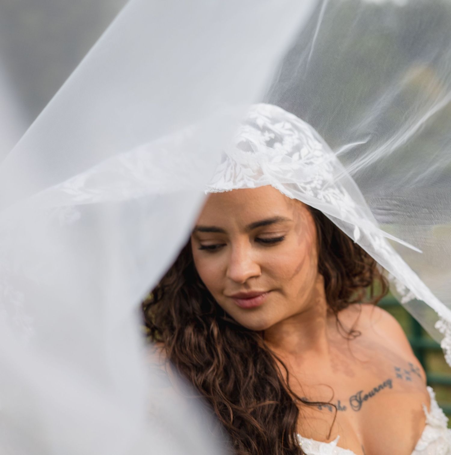 Close-up portrait of bride with curly hair and shoulder tattoo draped under a flowing lace veil.
