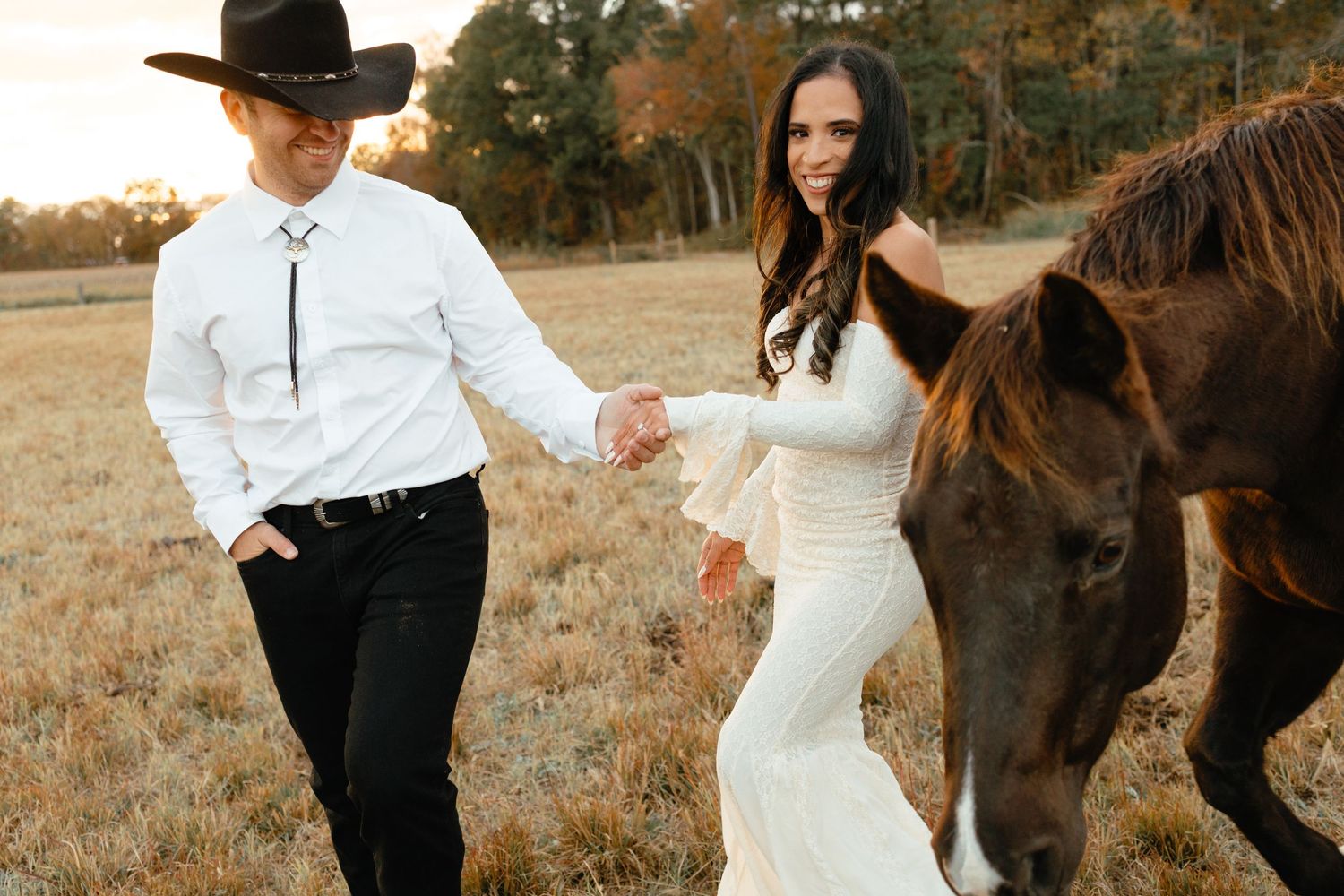 Bride and groom walk hand-in-hand with a brown horse through a sunlit autumn field in western wedding attire.